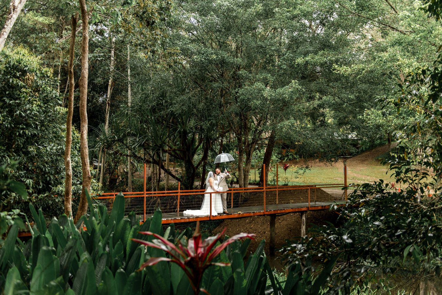Bride and groom share a romantic moment on a red bridge surrounded by lush tropical greenery.