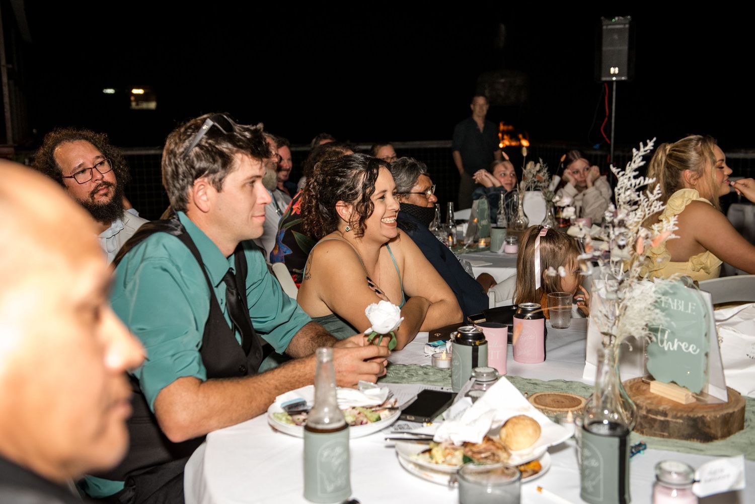 Wedding reception guests seated at round tables enjoying dinner under string lights at night.
