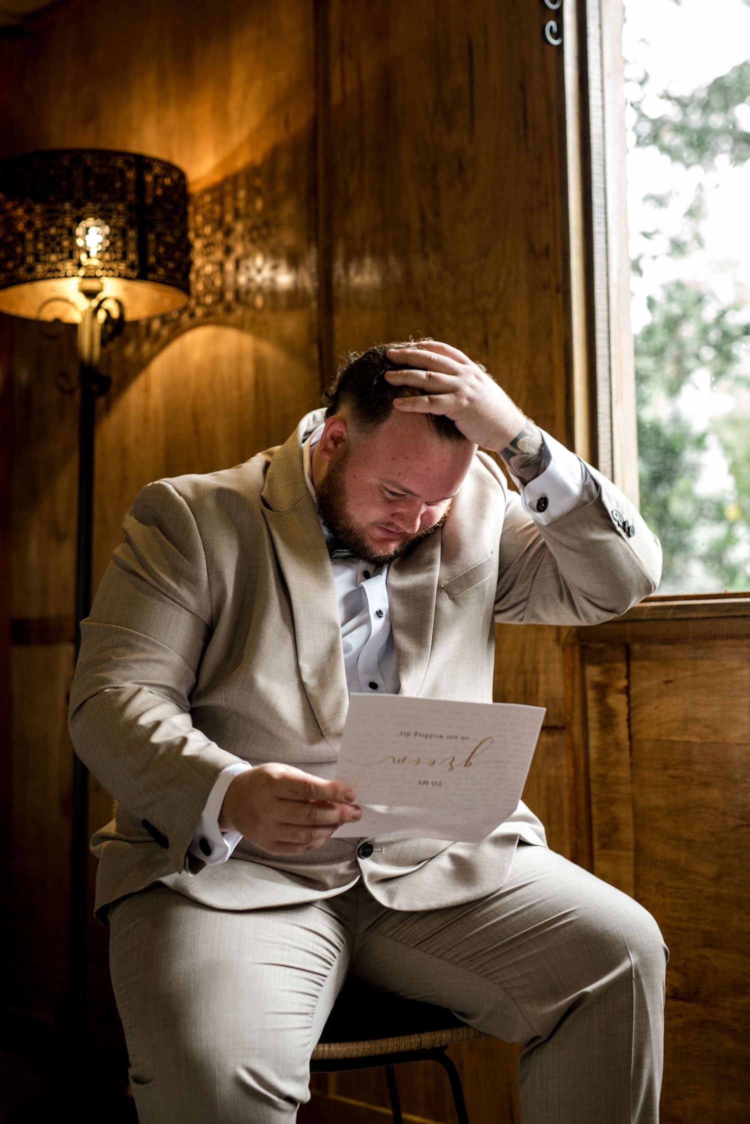 Emotional groom in beige suit reading heartfelt letter seated by window on wedding day.