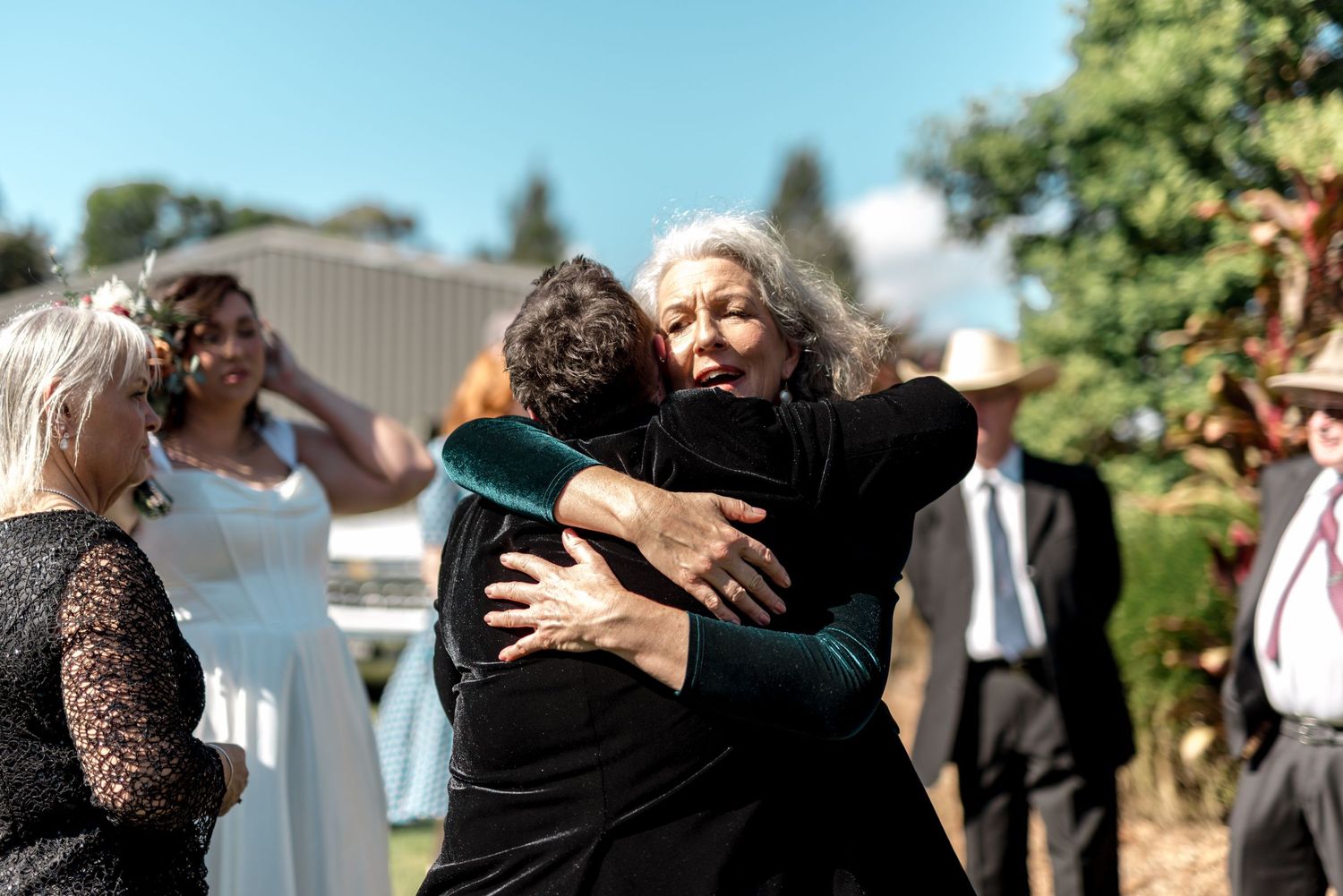 Two women share warm embrace at outdoor wedding celebration with bridal party in background.