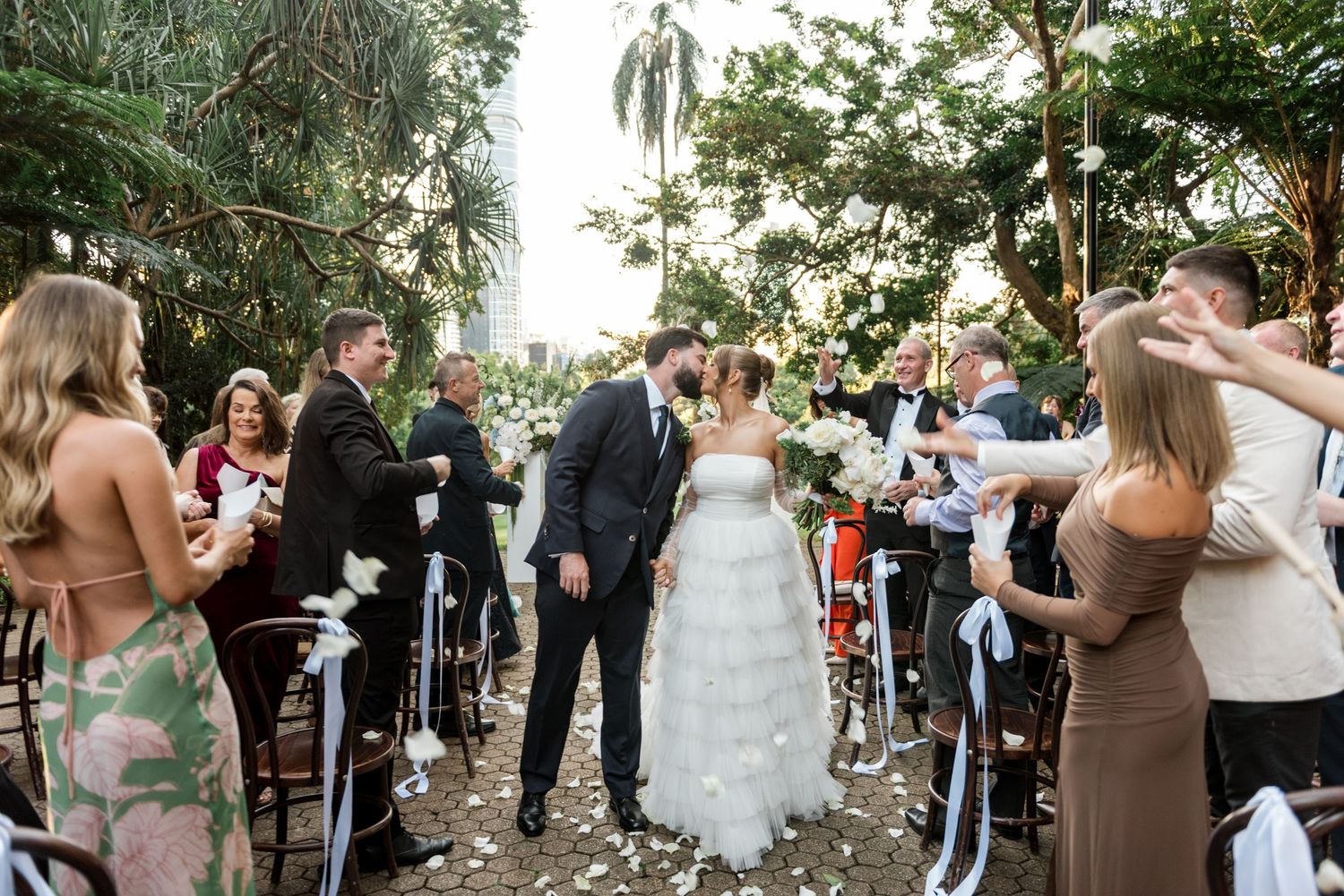 Bride and groom walk down the aisle as guests toss flower petals at outdoor garden wedding ceremony.