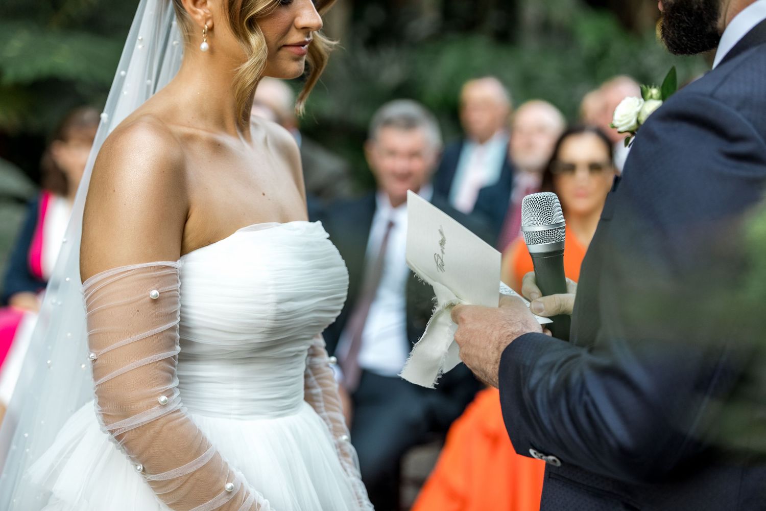 Bride in strapless white gown listens as groom reads vows into microphone at outdoor ceremony.