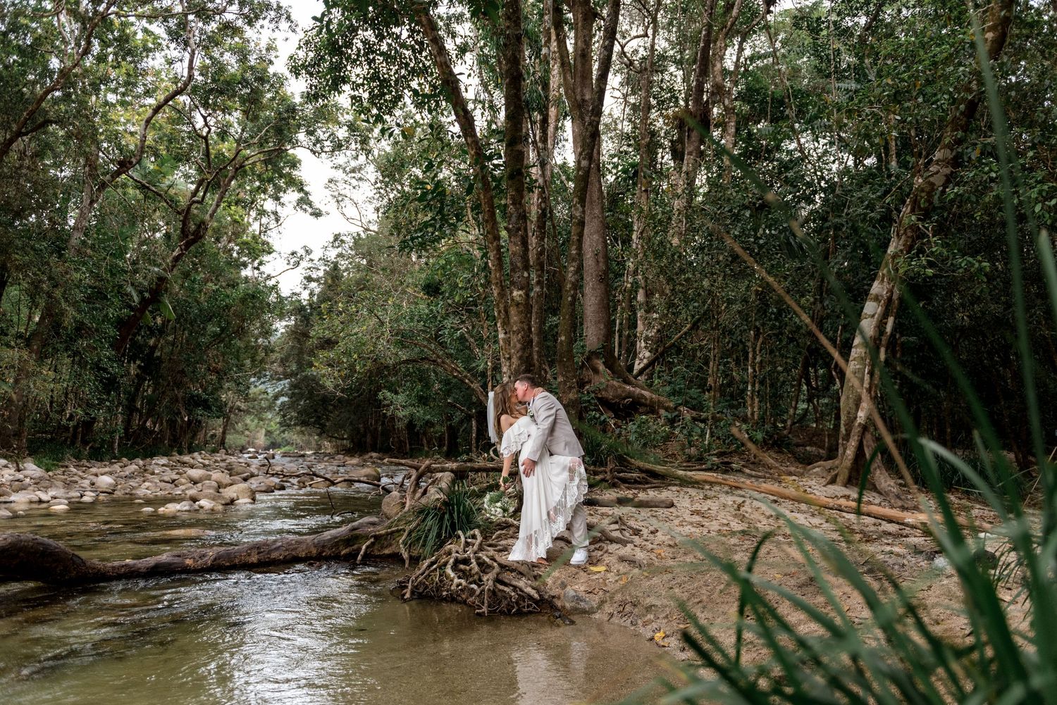 Bride and groom share a romantic moment on a fallen log over a serene forest creek.