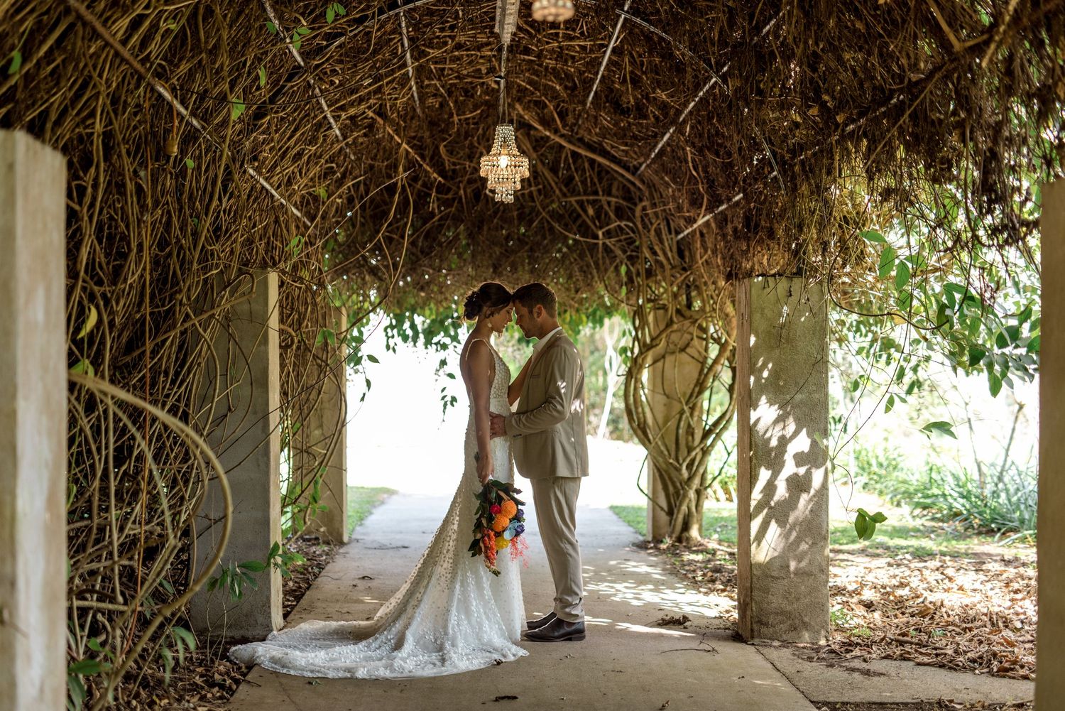 Bride and groom share an intimate kiss under a dramatic vine-covered pergola with chandelier.