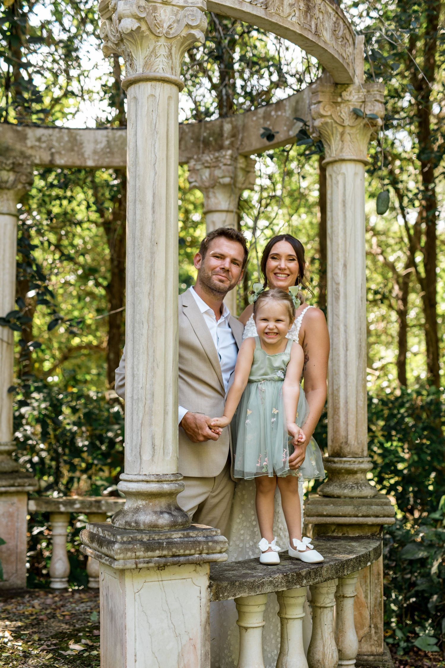 Smiling family of three posing on stone balustrade of classical garden gazebo surrounded by green trees.