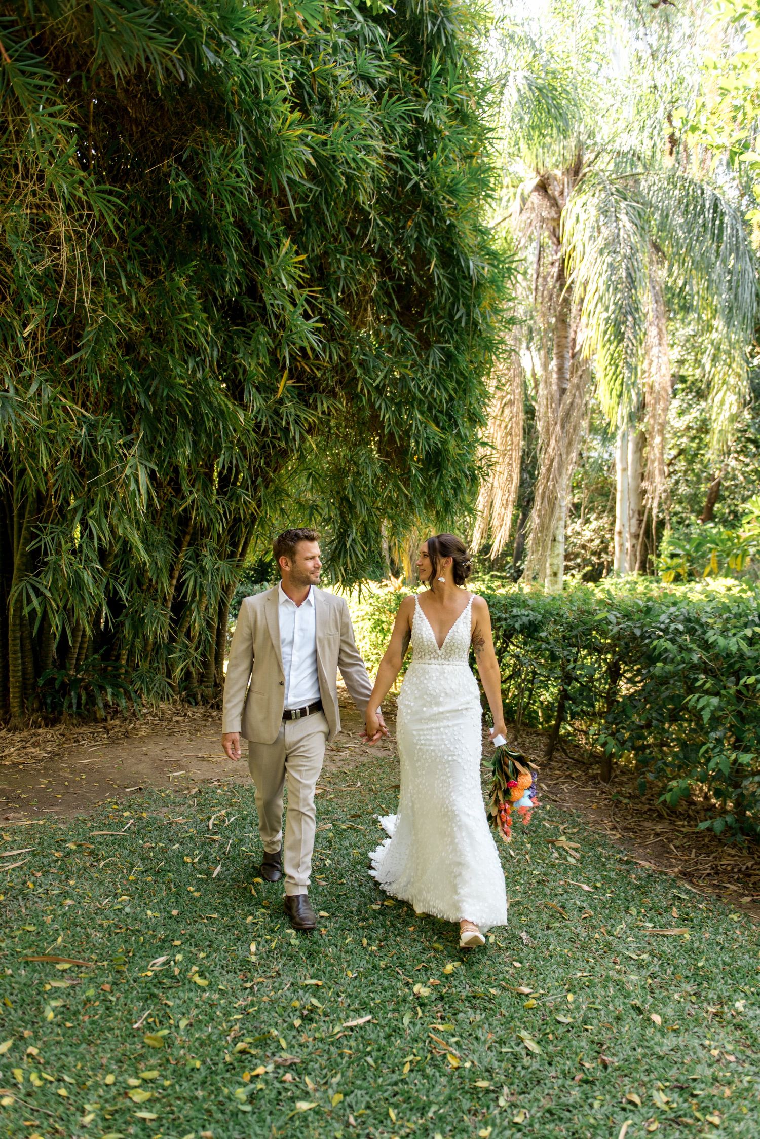 Bride and groom walking hand in hand through a lush green garden path on their wedding day.