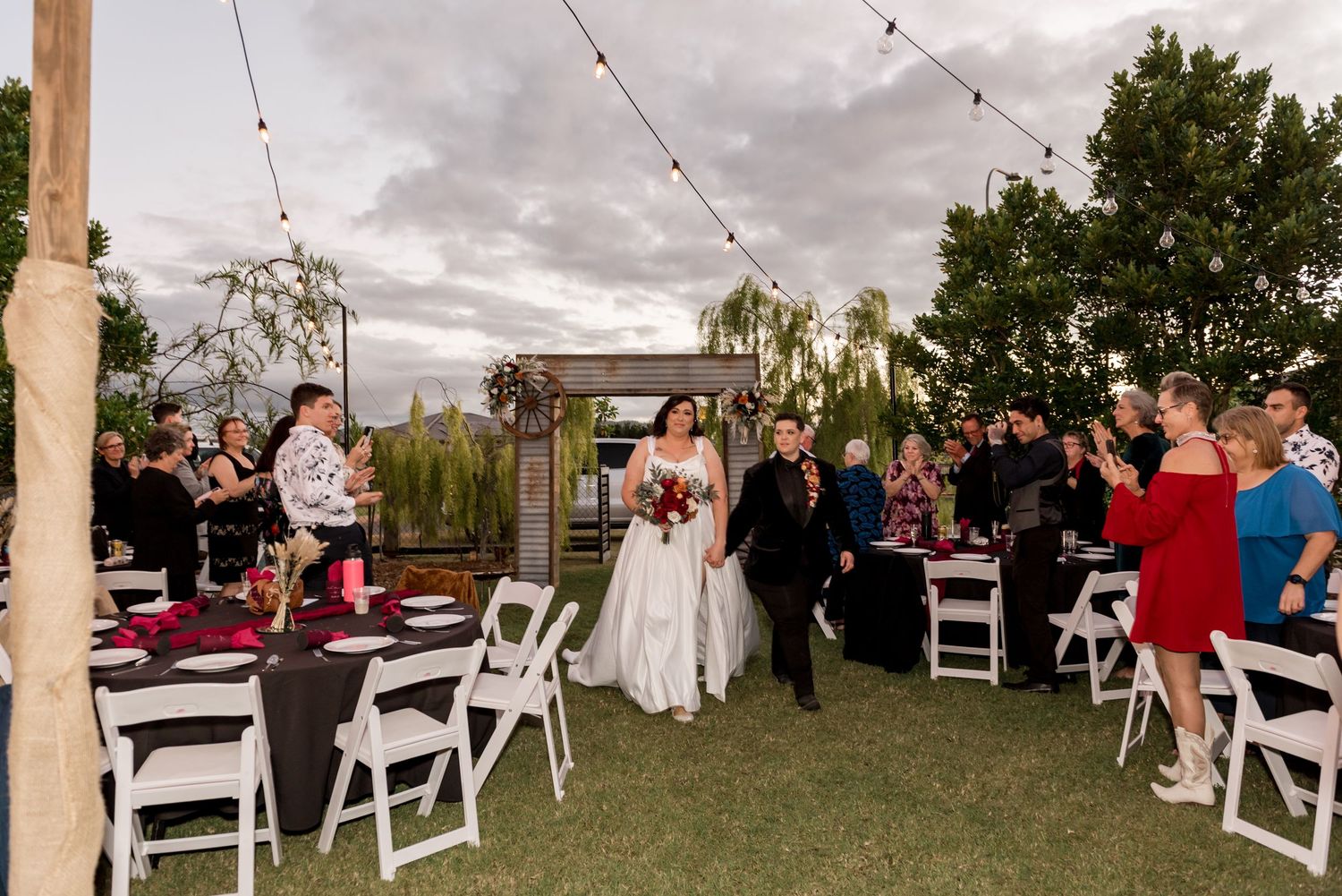 Bride and groom walk through guests at outdoor wedding reception with string lights overhead.