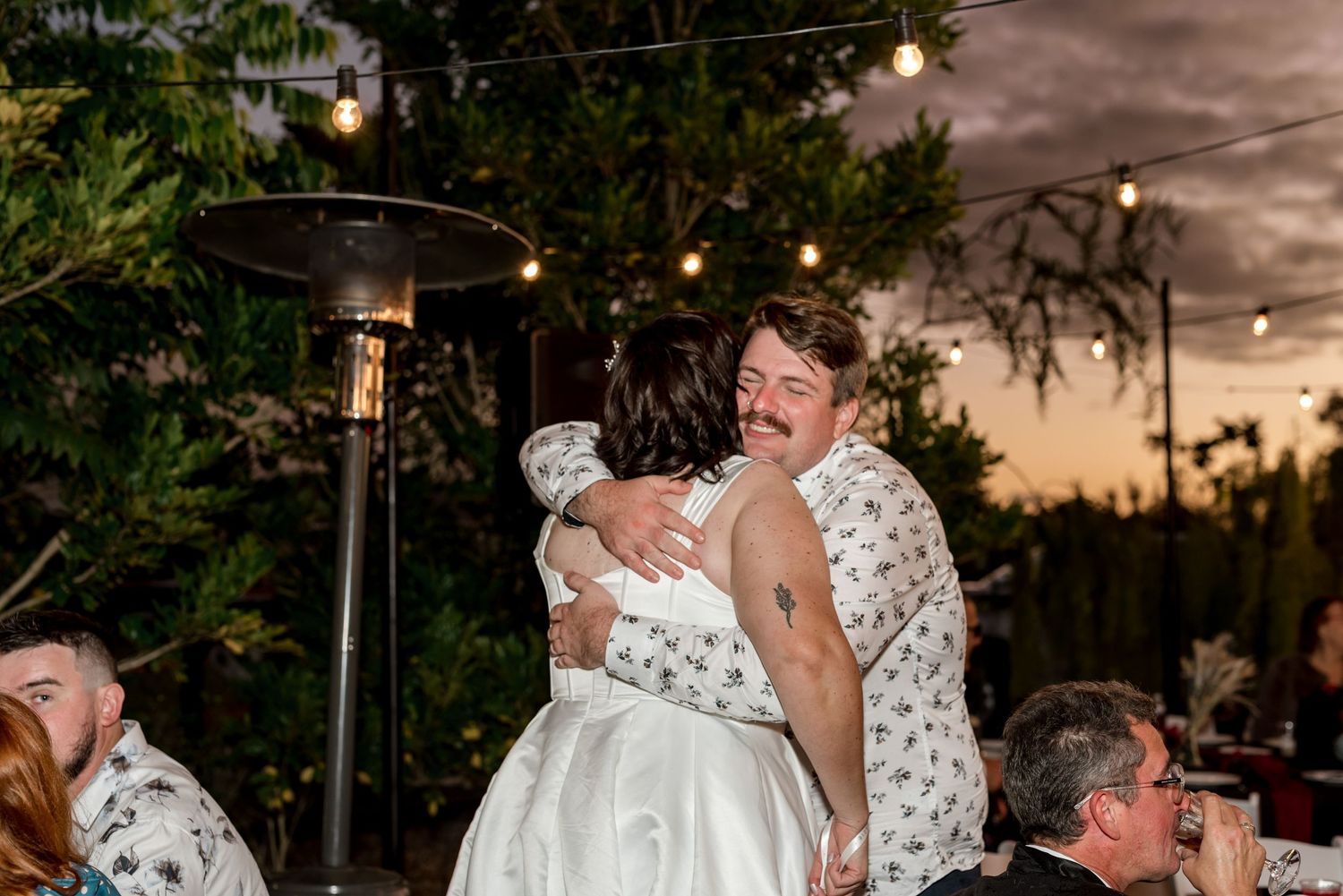 Couple sharing a warm hug at an outdoor wedding reception at dusk.