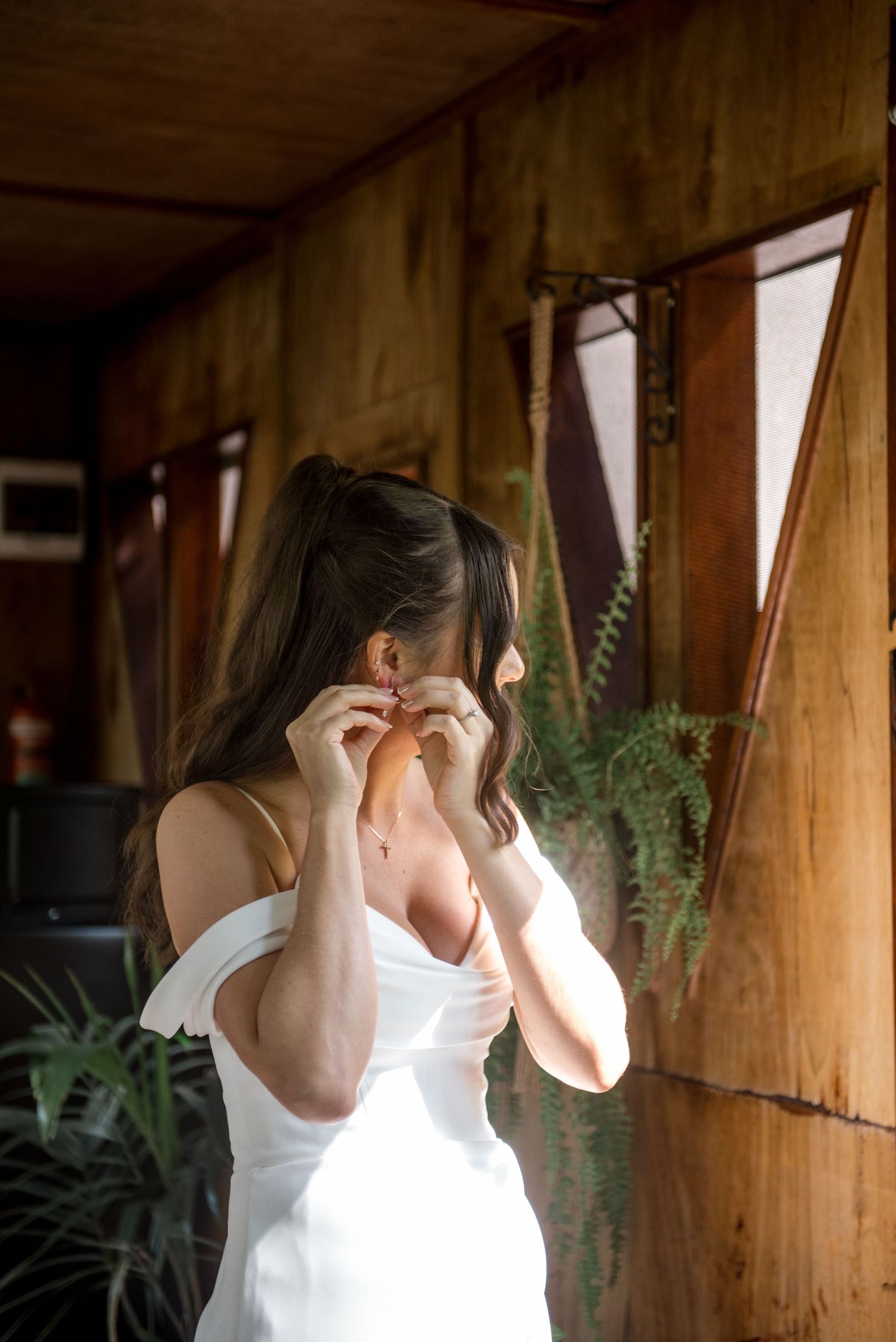 Bride in off-shoulder white dress putting on earrings inside a rustic wooden room.