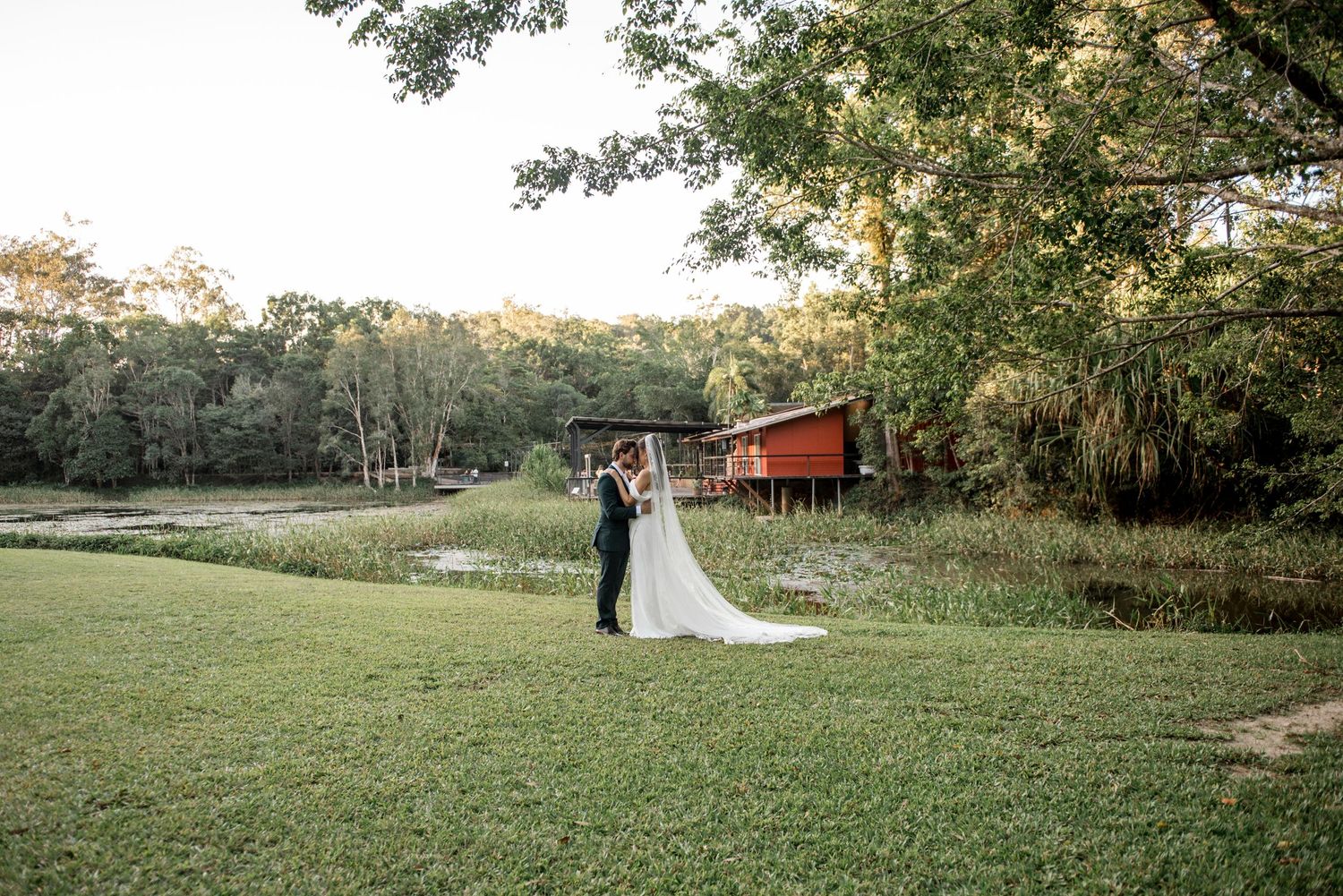 Bride and groom share a romantic moment on green lawn beside a serene pond at golden hour.