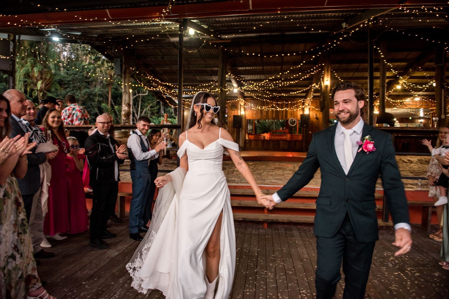 Bride and groom walk hand-in-hand through cheering guests at outdoor wedding reception.