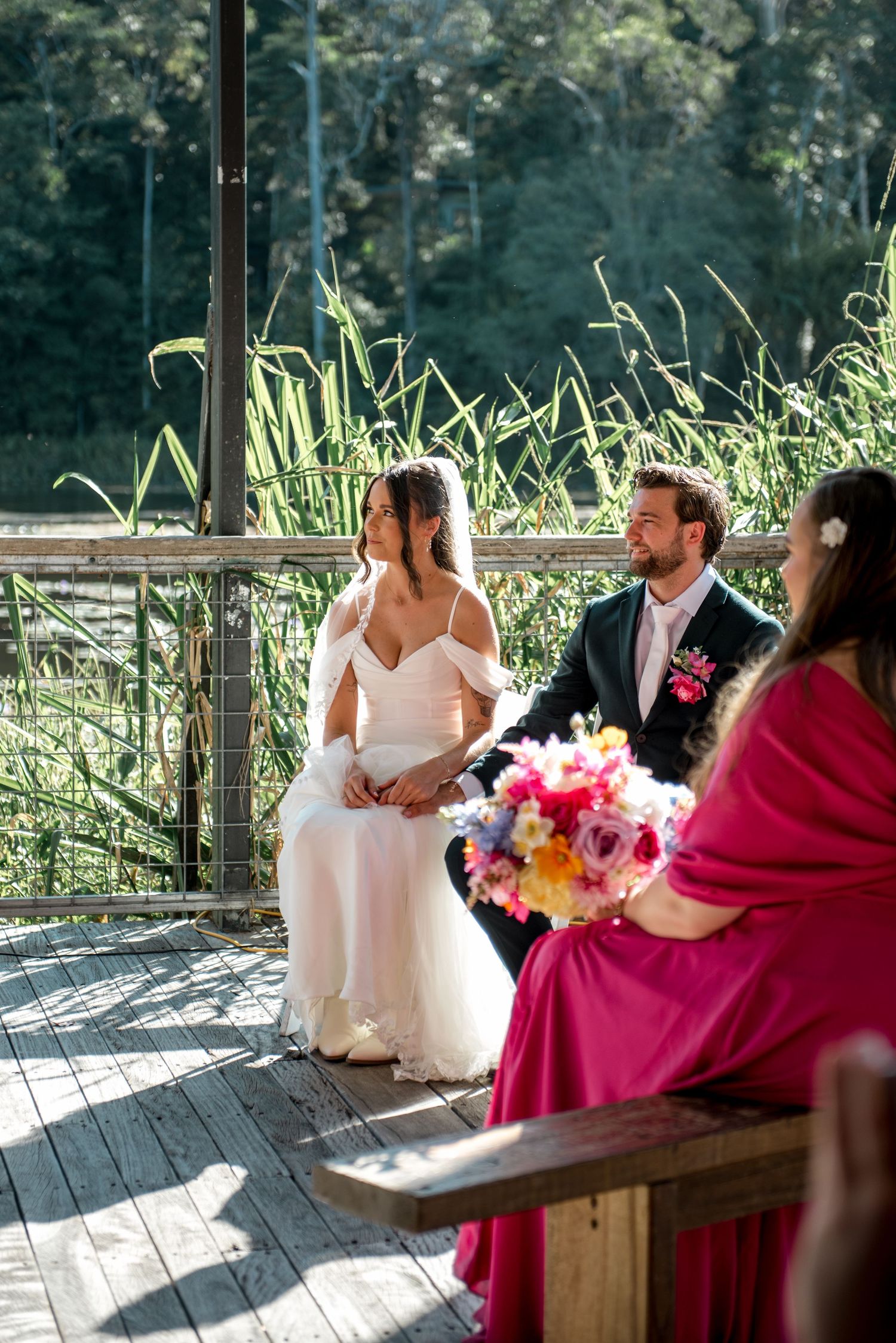 Bride and groom sit together on wooden deck during ceremony with lush green reeds in background.