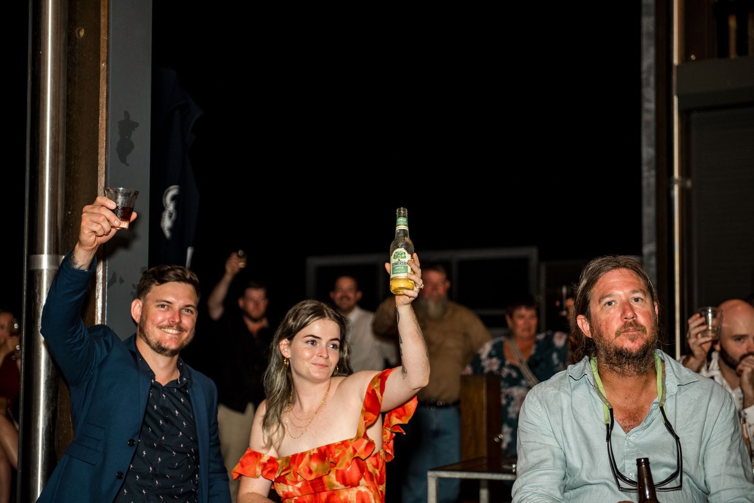 Three wedding guests raise drinks in a toast during an outdoor evening wedding reception celebration.