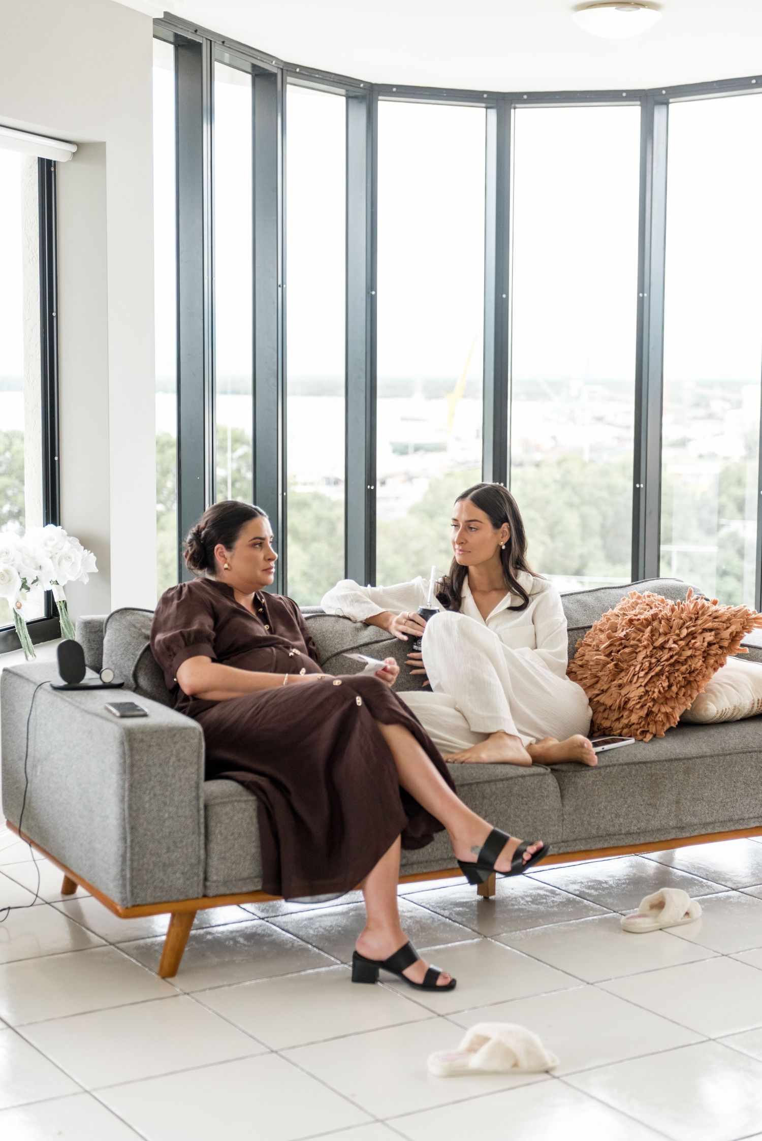 Two women in silk pajamas sit on a modern grey sofa chatting in a bright high-rise apartment with panoramic windows.