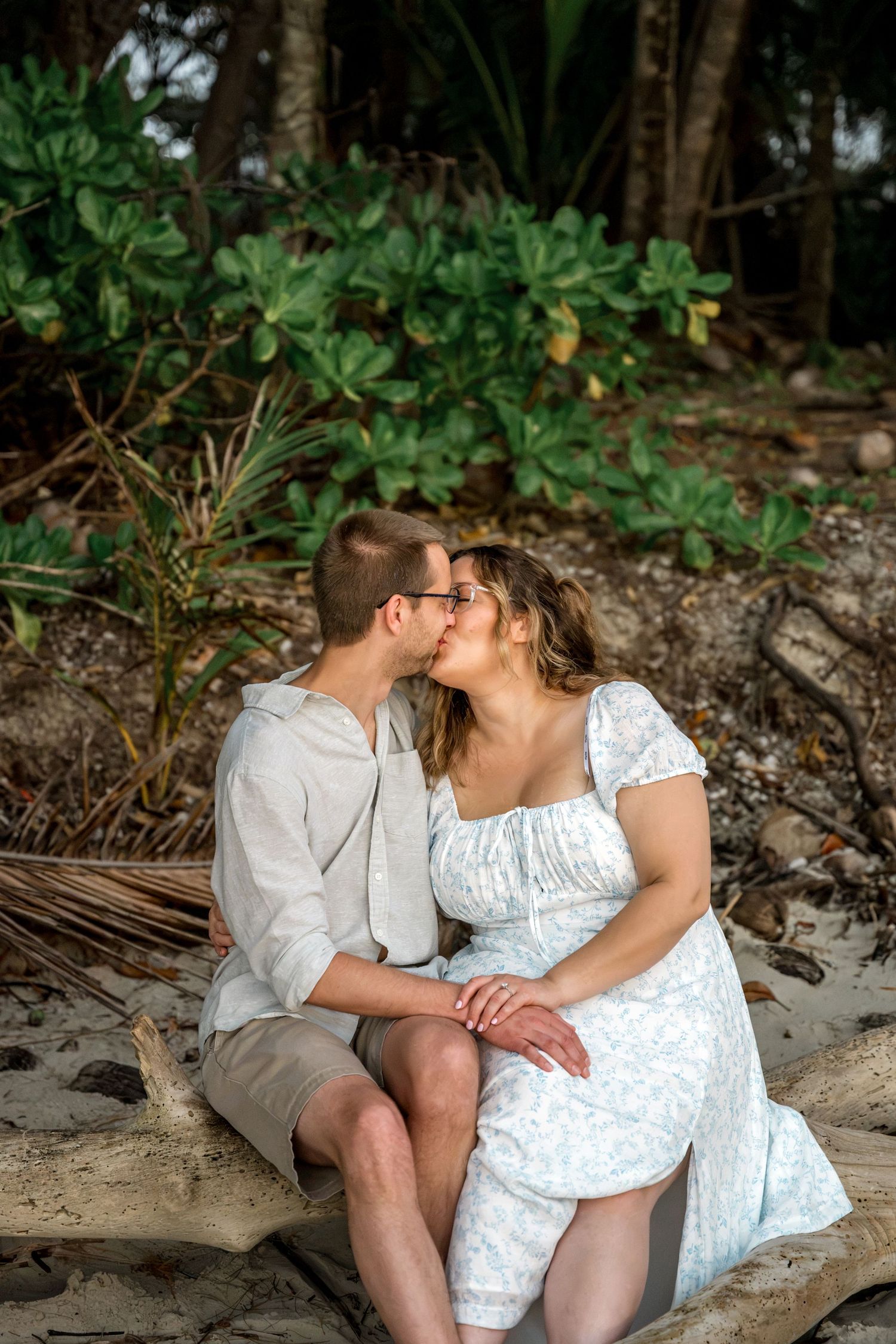 Couple sharing a romantic kiss while sitting on driftwood on a tropical beach surrounded by lush greenery at Four Mile Beach, Port Douglas.