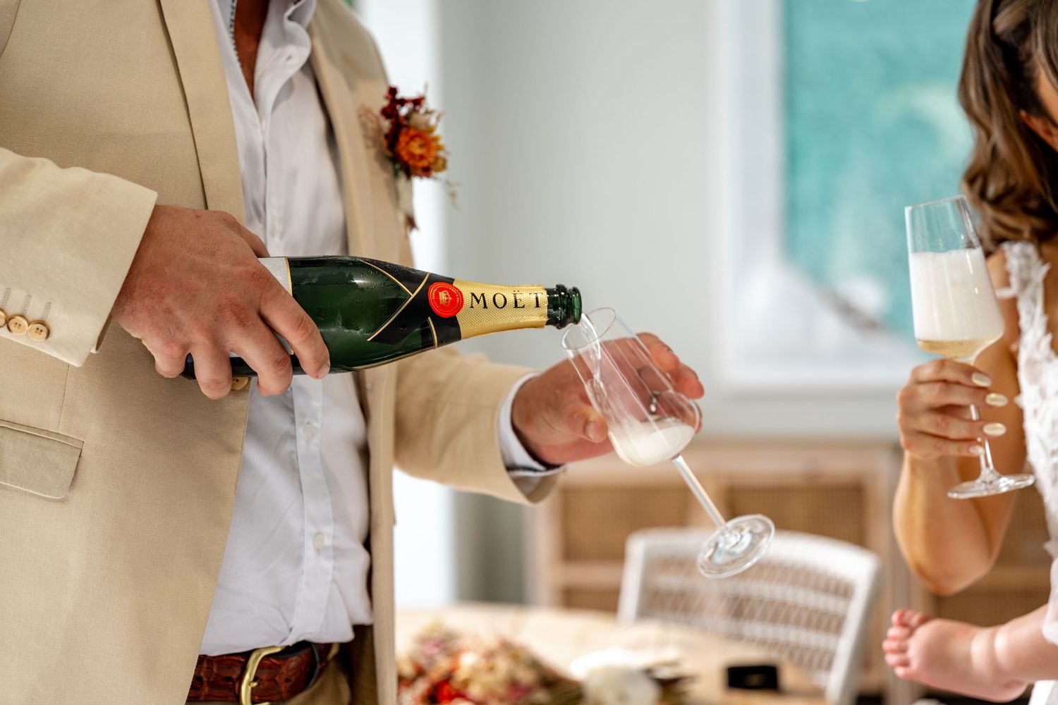 Groom in beige suit pouring Moet champagne into glass at elegant wedding reception table setting.