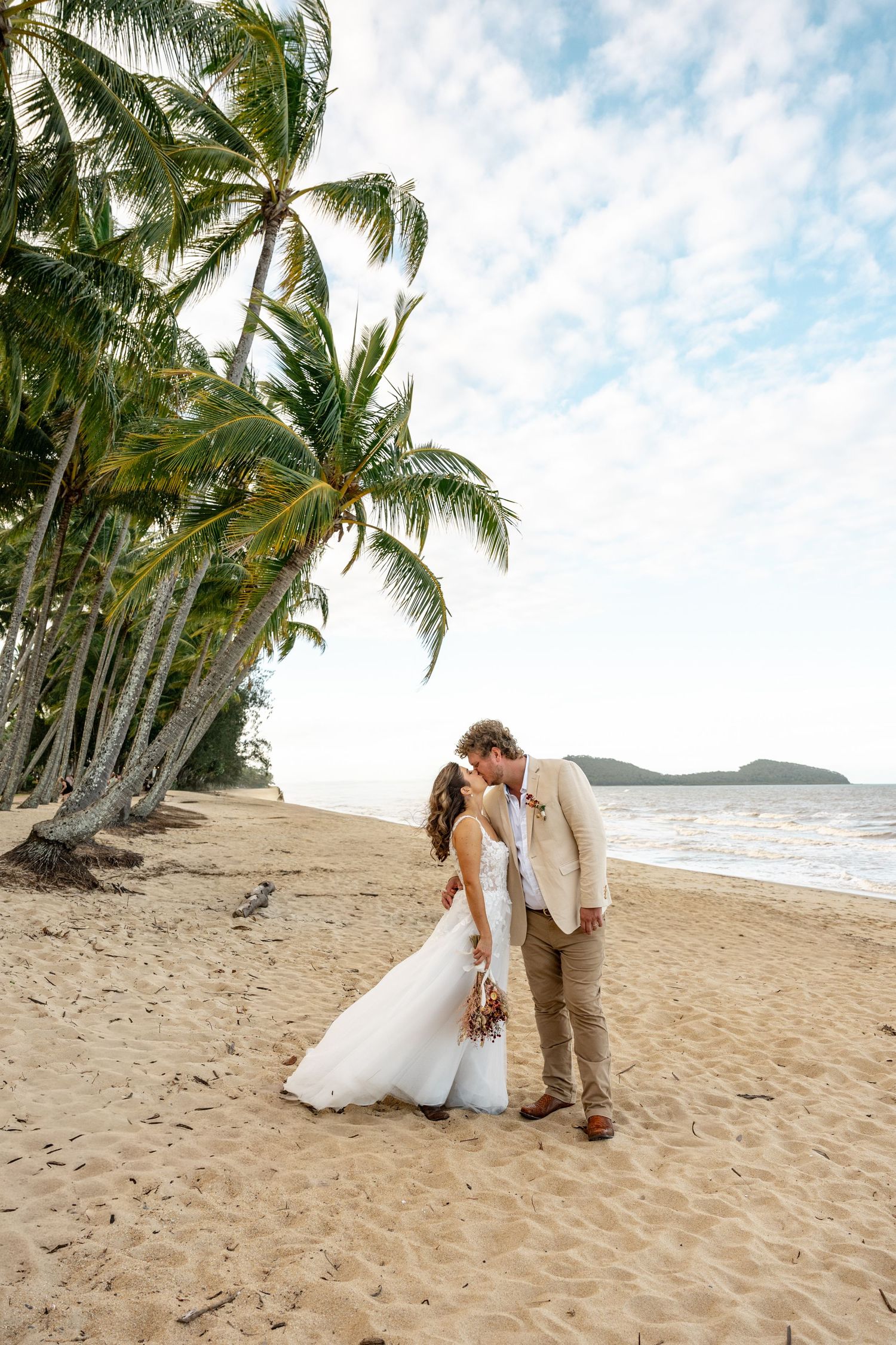 Bride and groom share a tender kiss on a palm-lined tropical beach with turquoise ocean behind them.