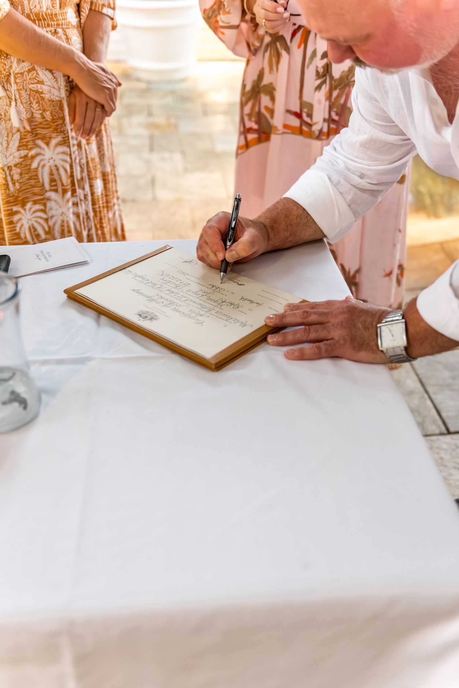 Groom signing official wedding certificate at white draped table during outdoor wedding ceremony.