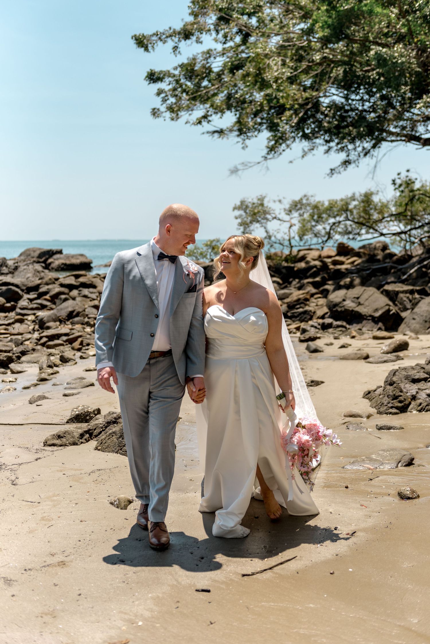 Smiling bride and groom walking hand in hand on a sunny beach with rocks and ocean views.