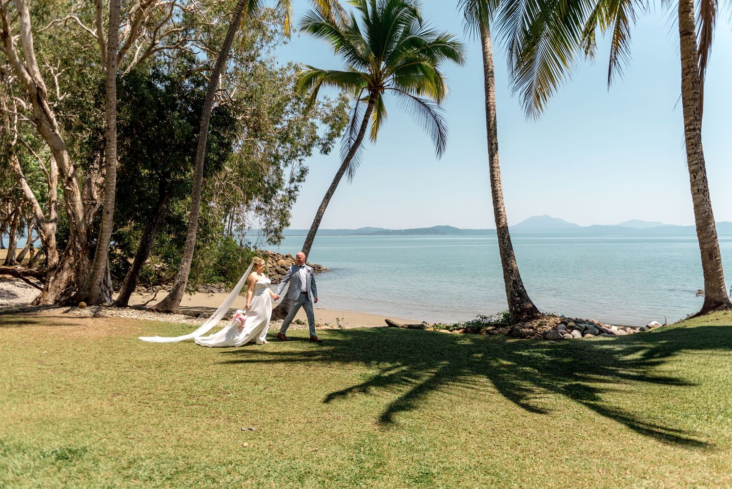 Groom twirls his bride in a flowing white gown on a sunny tropical beachfront lawn with palm trees.