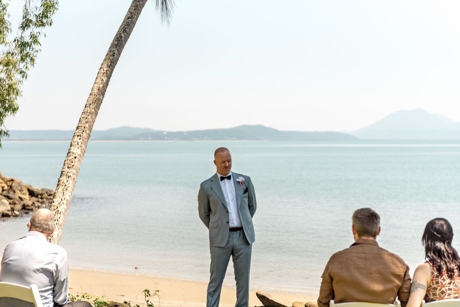 Groom in gray suit and bow tie awaits bride at outdoor beach wedding ceremony.