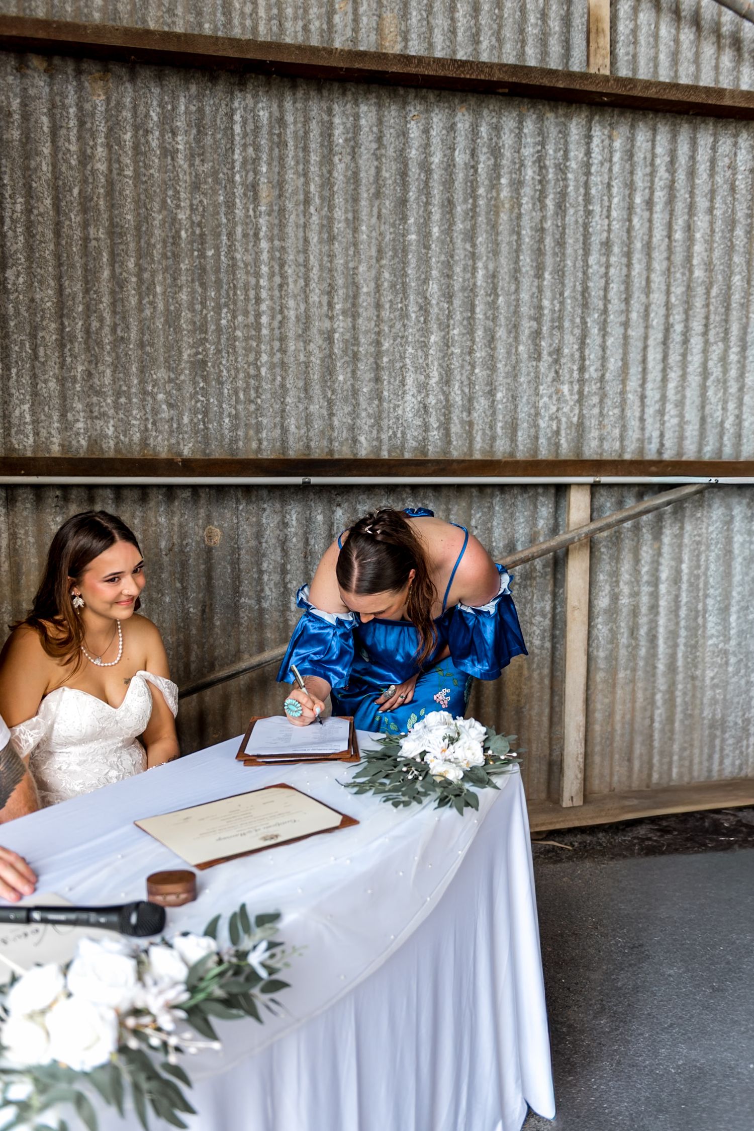 Bridesmaid in blue dress signs wedding certificate while bride smiles nearby.