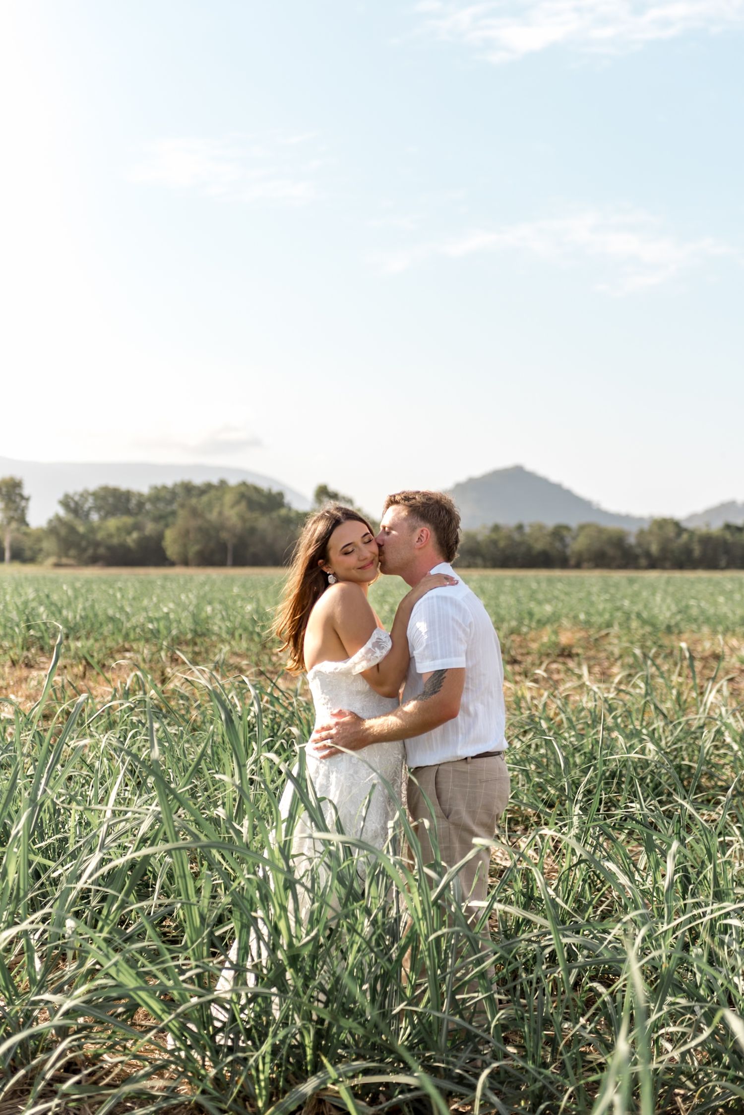 Man kissing woman on the cheek in a sunlit green field with a mountain backdrop.