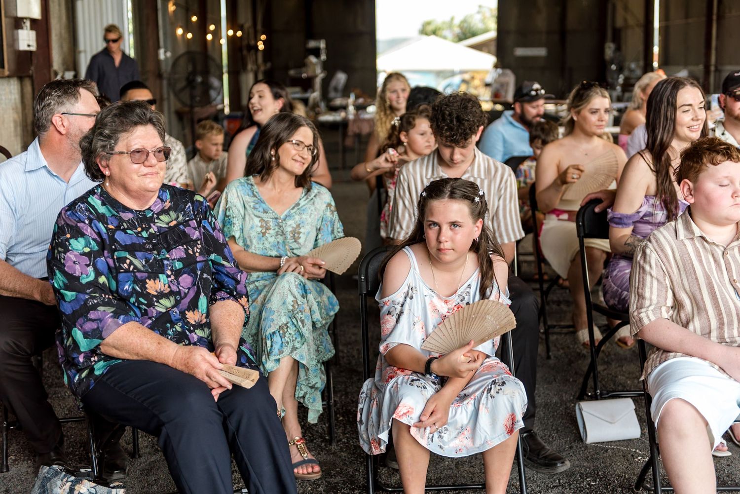 Smiling wedding guests seated in rows inside a barn venue, including a woman in a floral dress in the foreground.