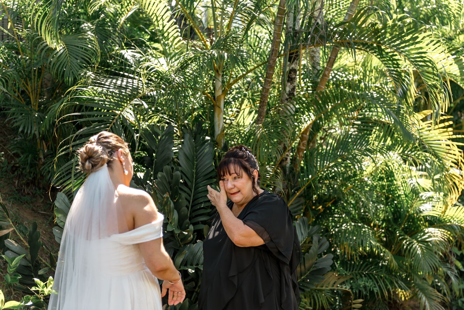 Emotional wedding guest wipes tears while bride in white veil stands in tropical garden.