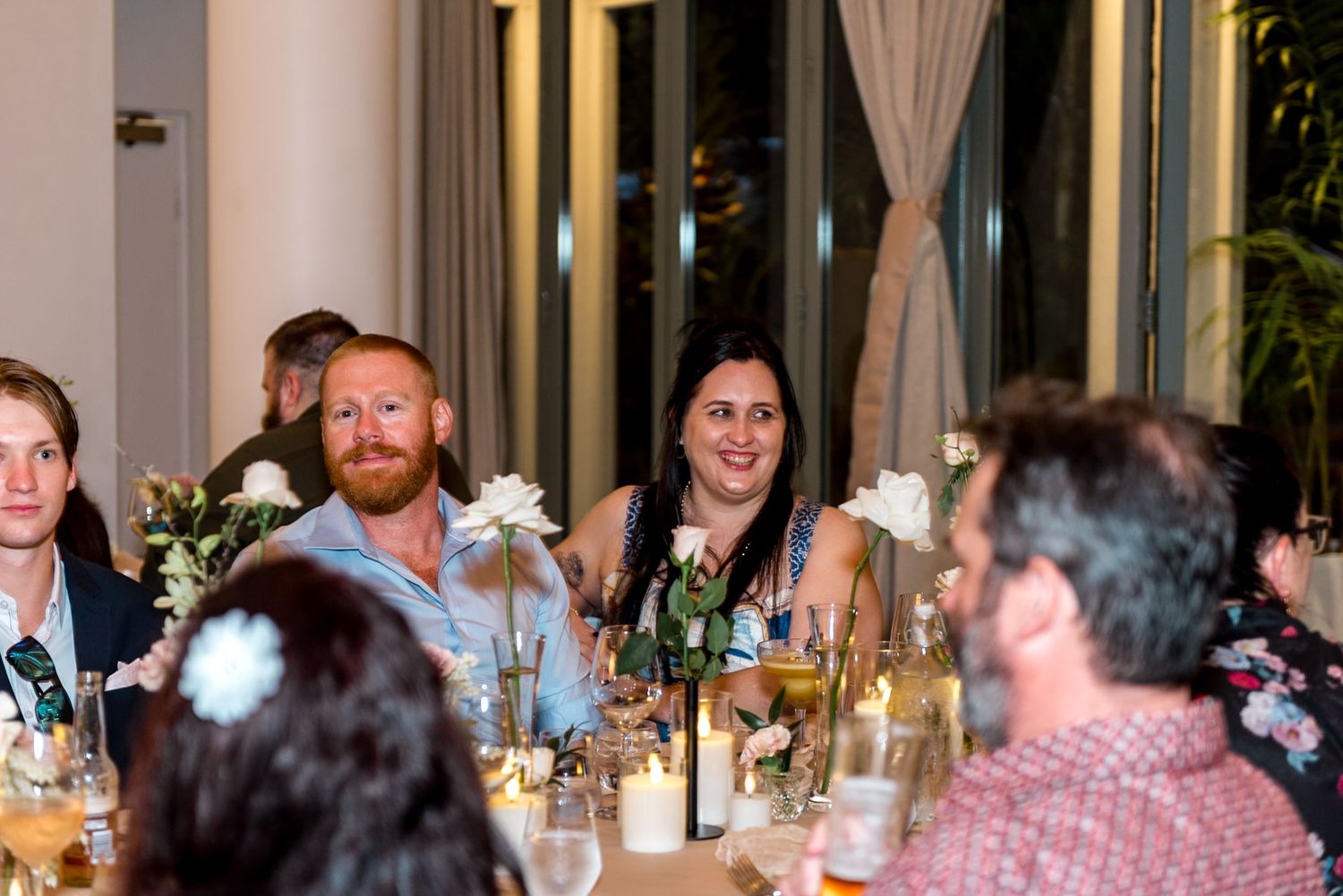 Smiling wedding guests enjoy dinner at candlelit reception table adorned with white floral arrangements.