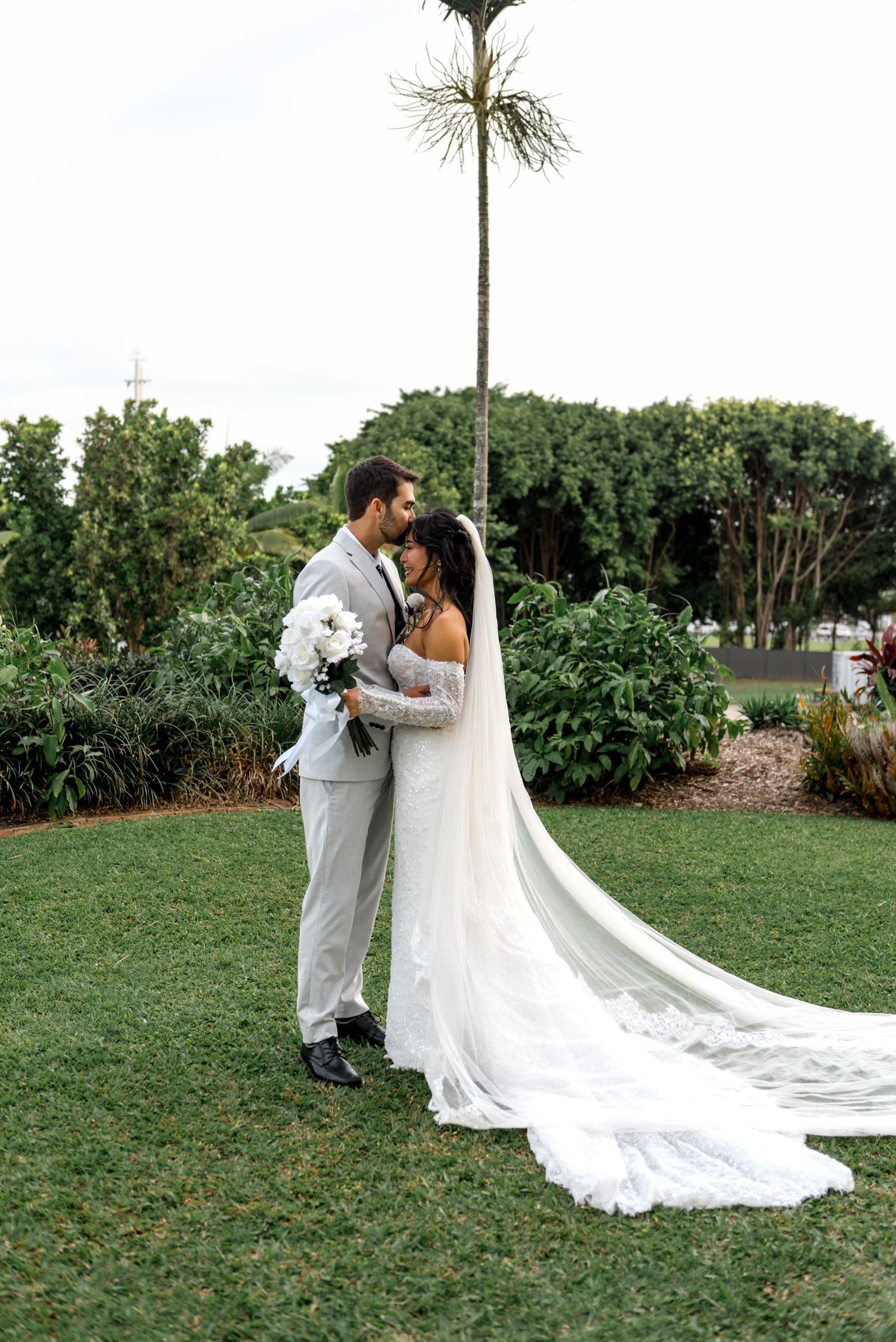 Bride and groom embrace on lush garden lawn with bride in lace gown and long cathedral veil.
