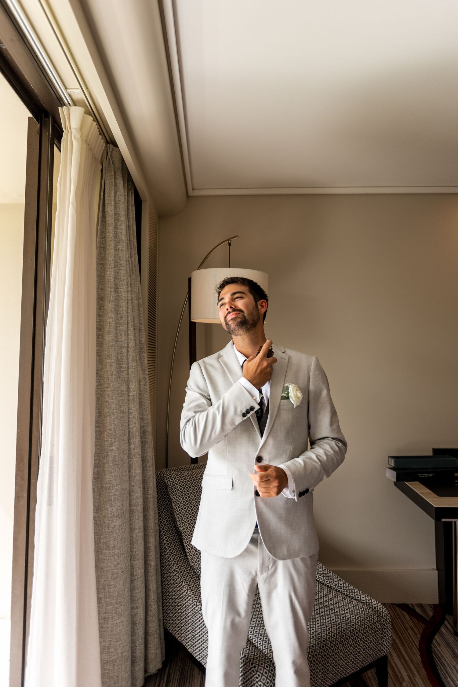 Smiling groom in light gray suit with floral boutonniere adjusting his tie near a window in a hotel room.