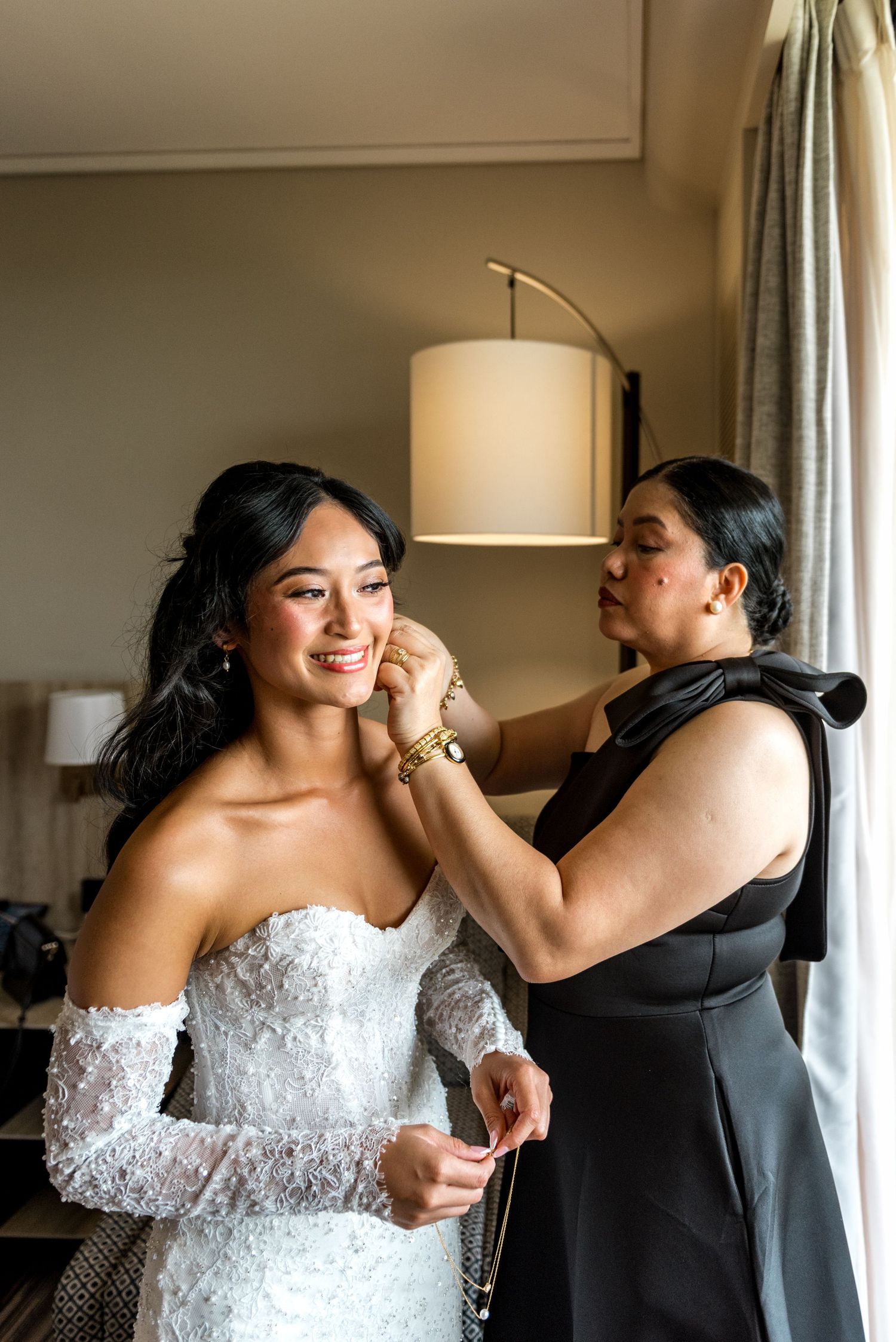Bride in off-shoulder lace wedding dress smiling as woman helps her with earrings.