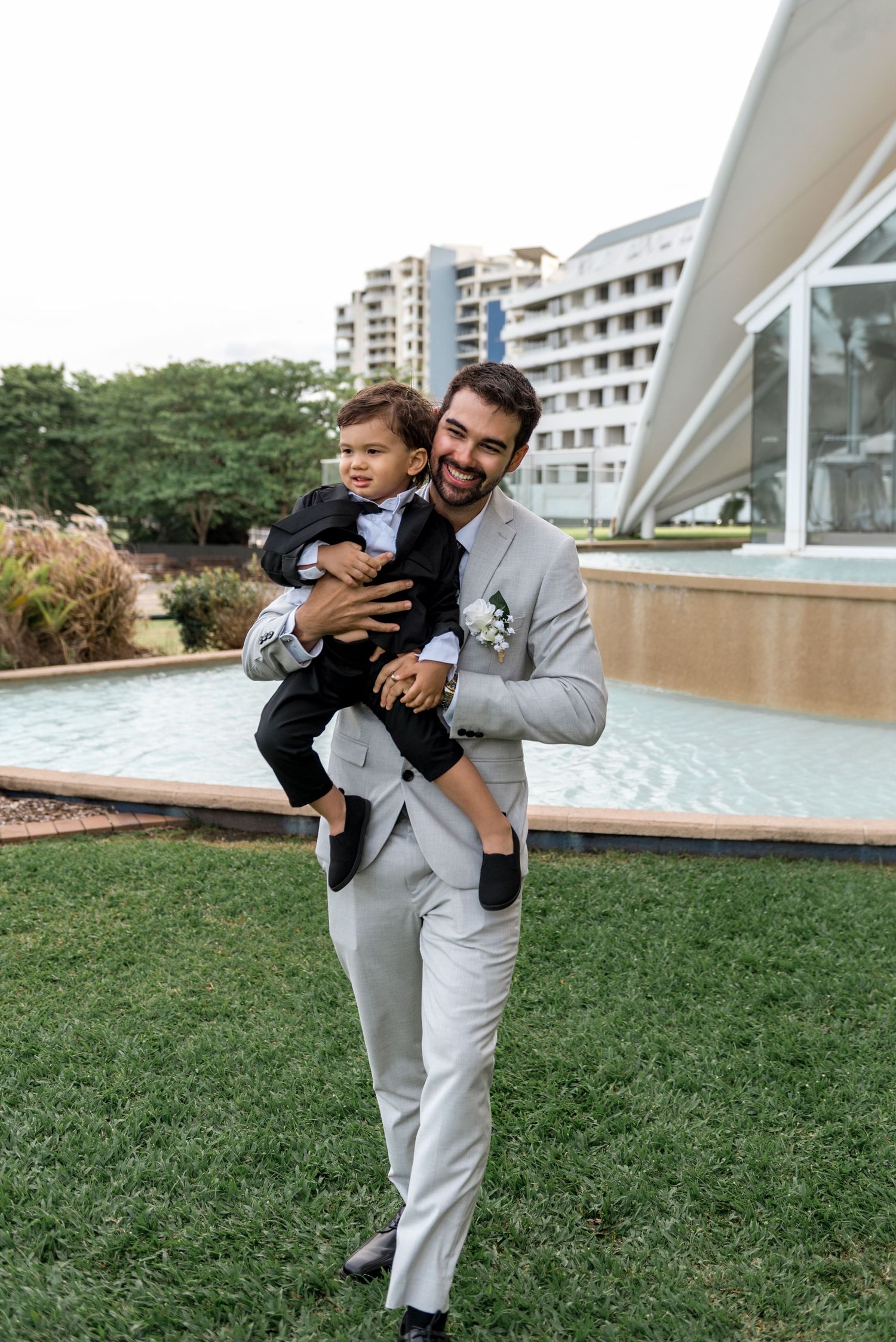 Smiling groom in light grey suit holding young child in black suit at outdoor wedding venue.