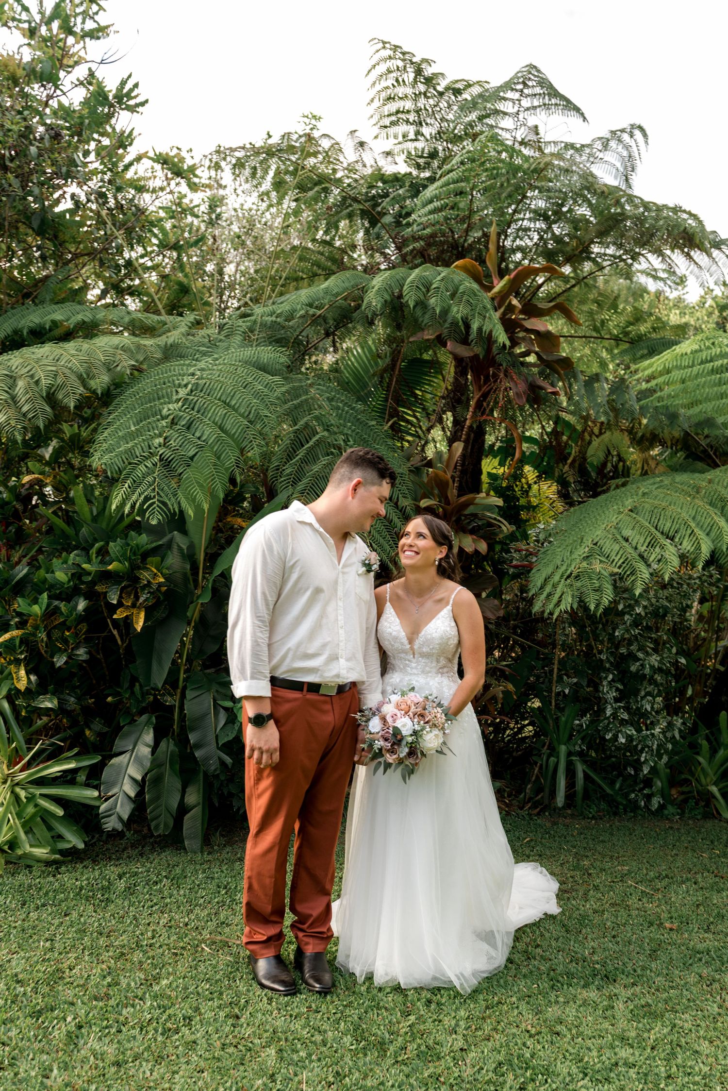 Bride and groom share a romantic moment in front of lush tropical ferns and greenery.