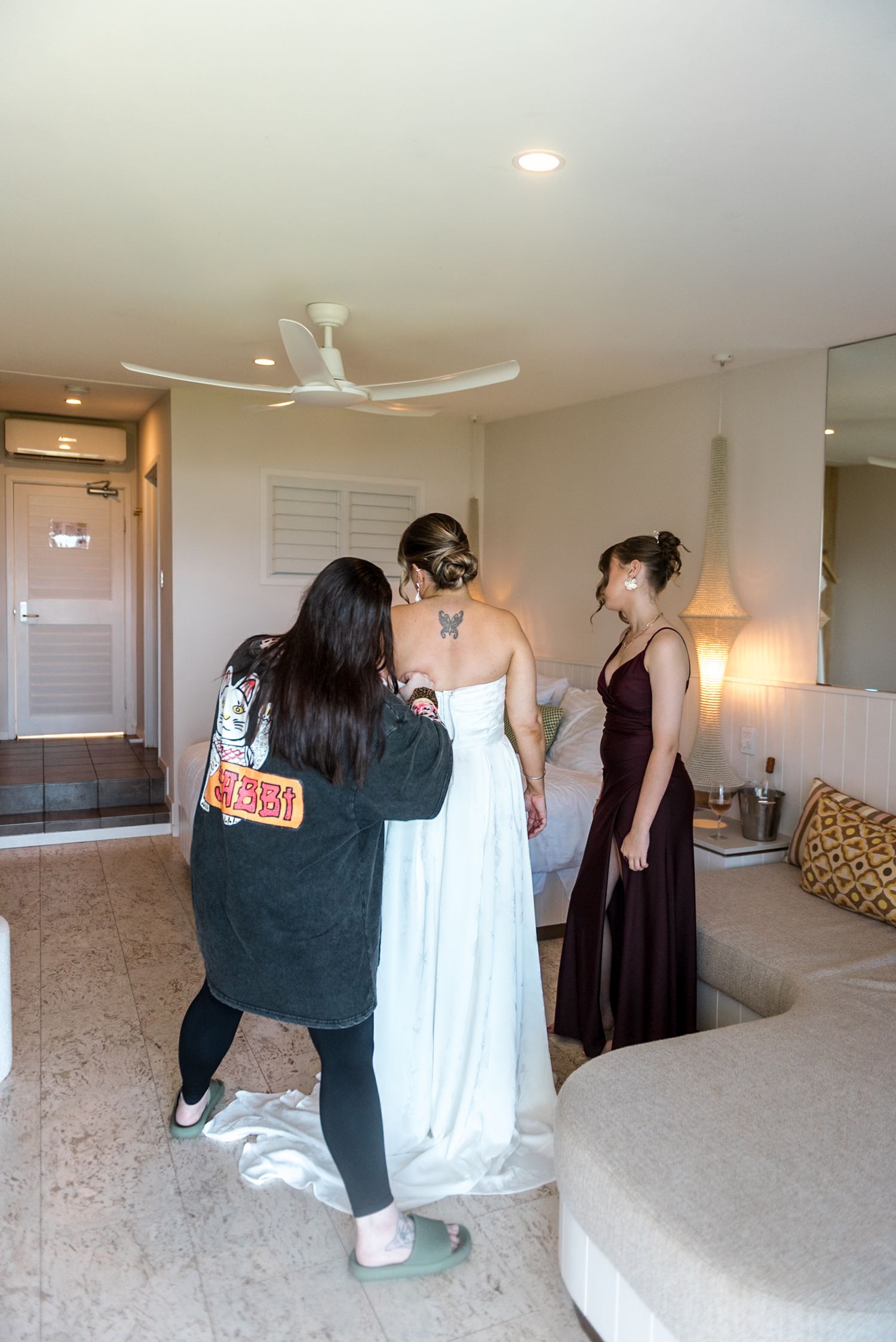 Bride in white gown being dressed by two women in a warmly lit hotel room.