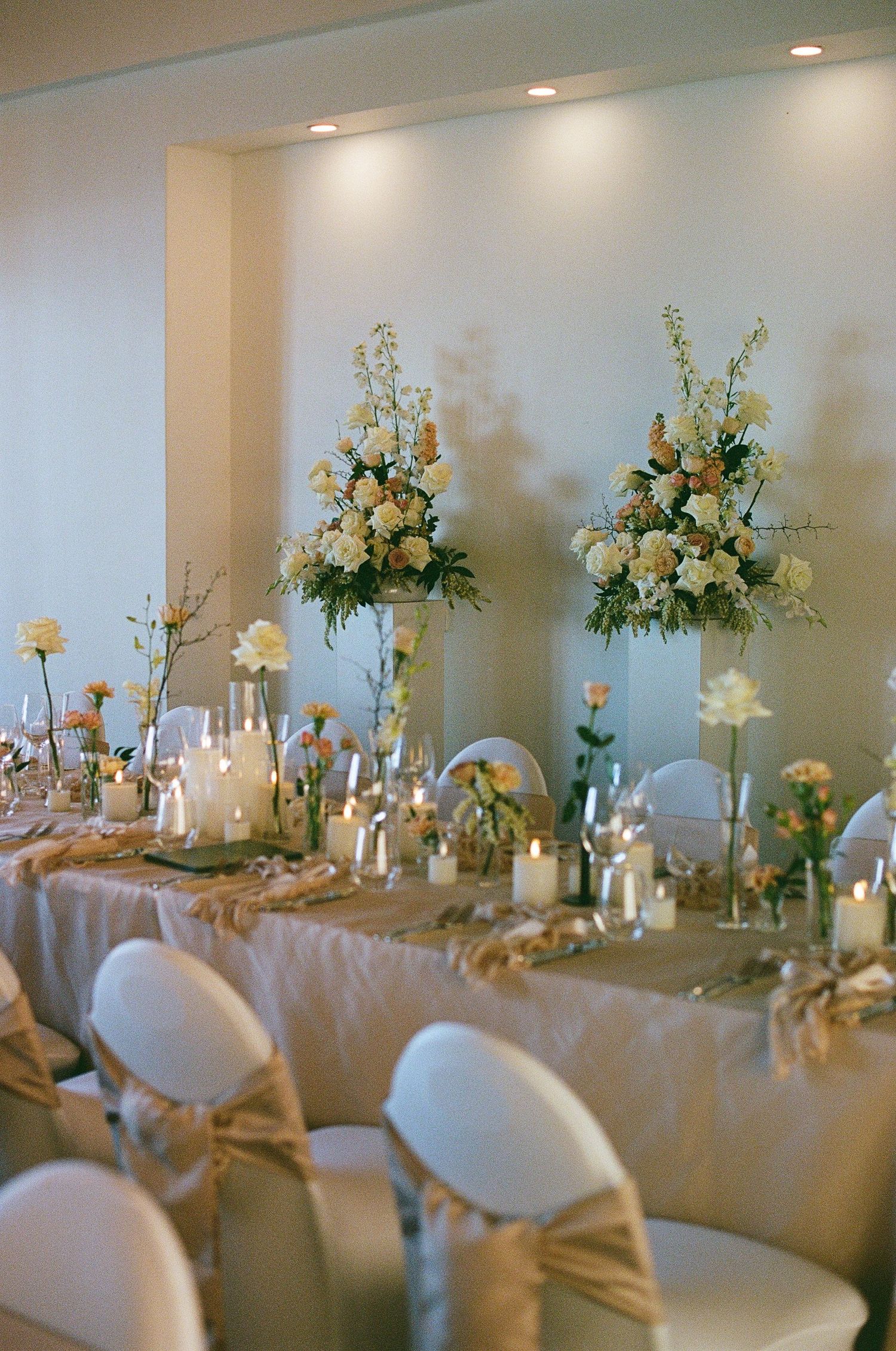 Wedding reception head table adorned with tall floral arrangements, candles, and neutral satin chair bows.