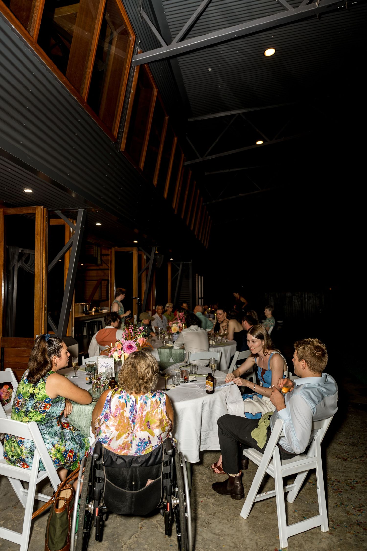 Guests including a wheelchair user dining at long tables during an outdoor evening reception.