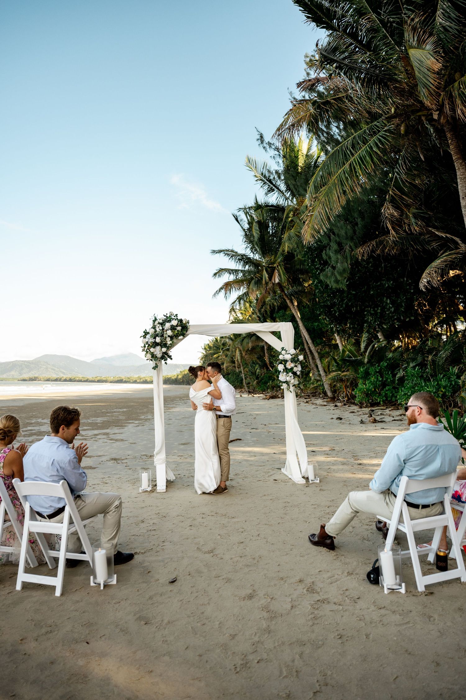Bride and groom share first kiss at a beach wedding ceremony with guests seated on white chairs at Four Mile Beach, Port Douglas.