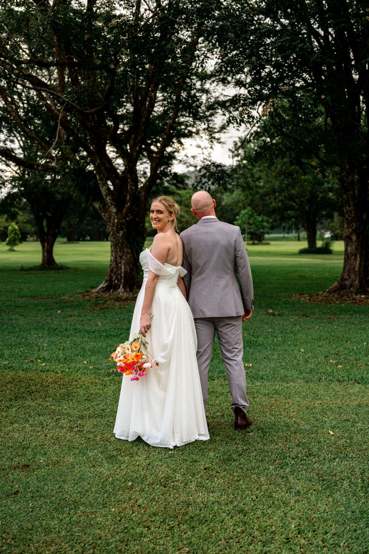 Bride in white gown and groom in grey suit walking away through tree-lined park, bride glancing back over shoulder.