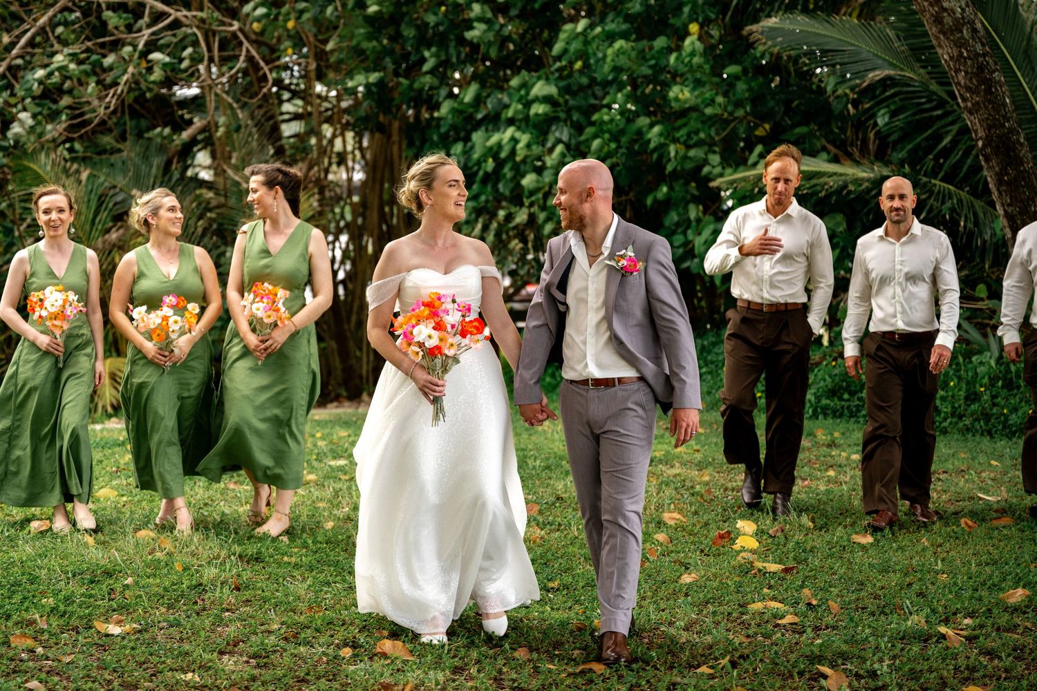 Wedding party walks through lush green garden, bride in white gown with bridesmaids in sage green dresses and groomsmen in brown pants.