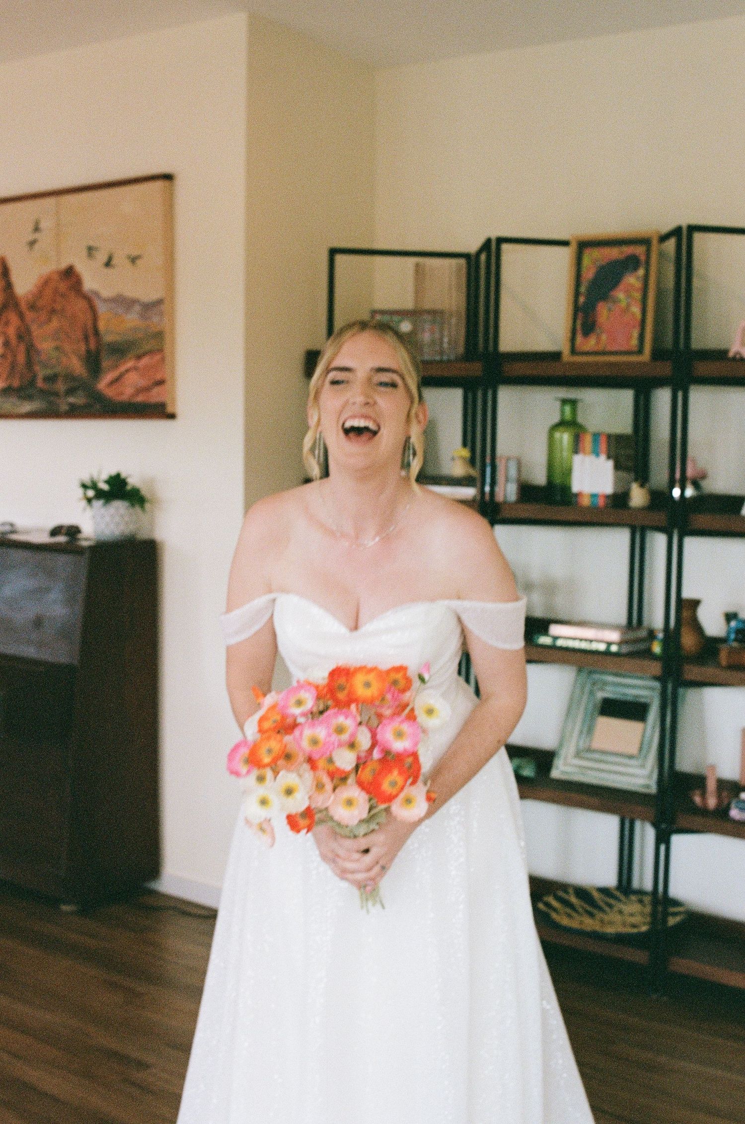 Laughing bride in white off-shoulder wedding dress holding vibrant orange and pink bouquet.