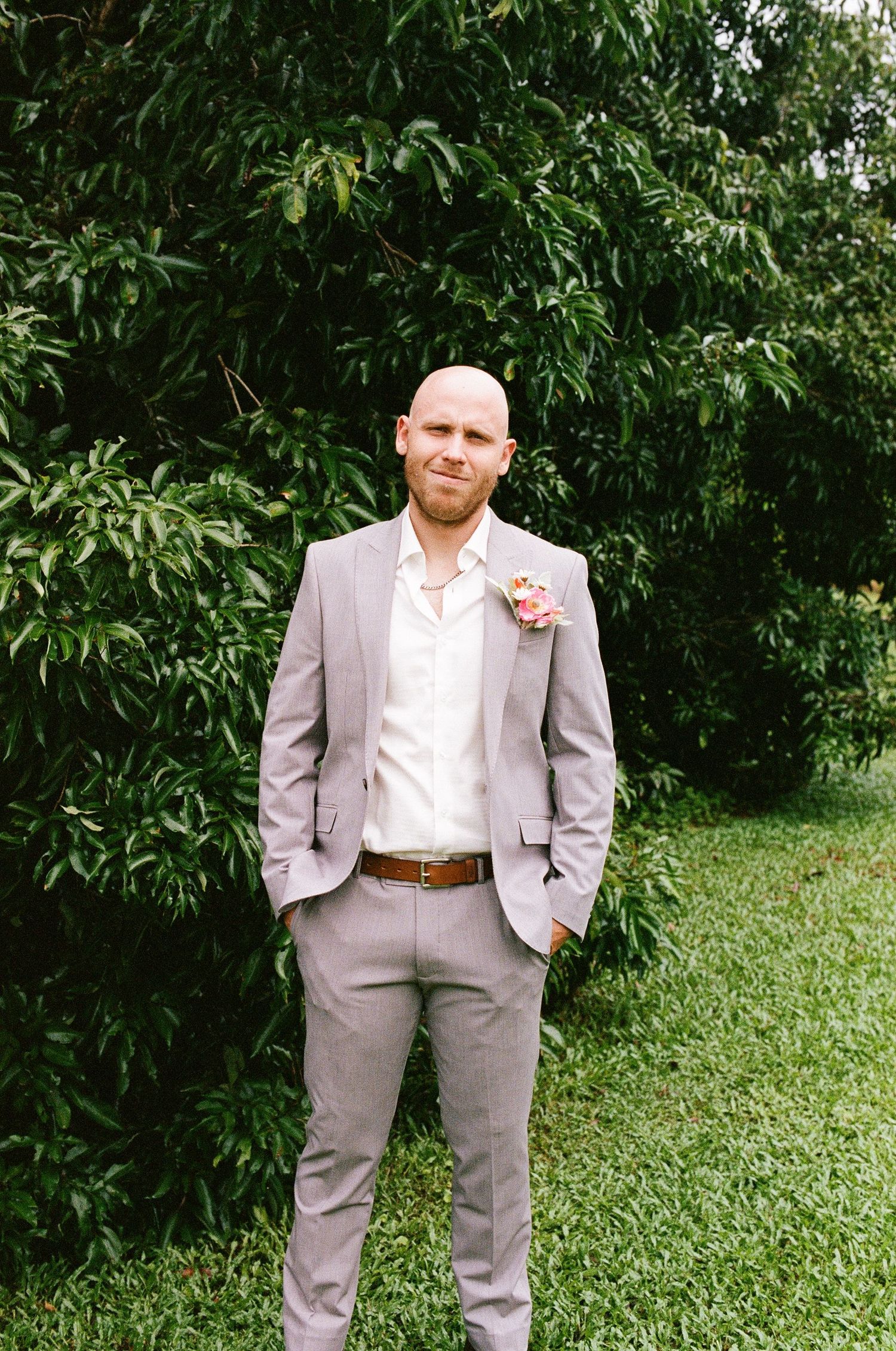Groom in a light gray suit with a floral boutonniere stands before lush green trees.