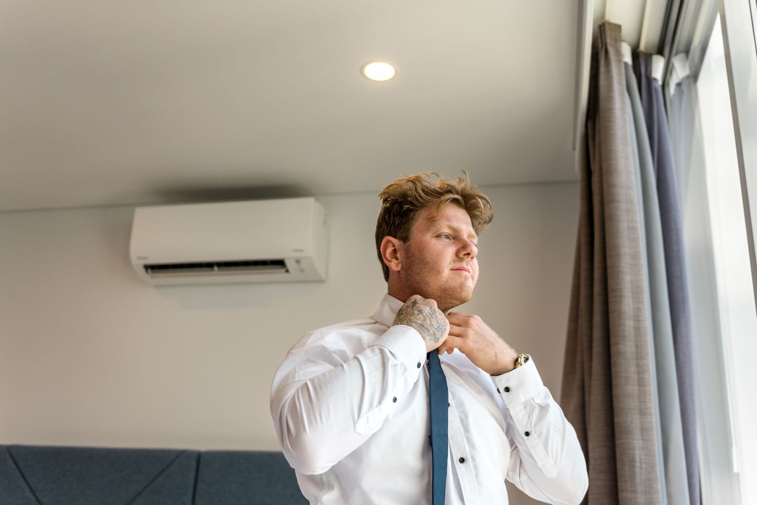 Groom adjusting blue tie while wearing white dress shirt in hotel room before wedding.