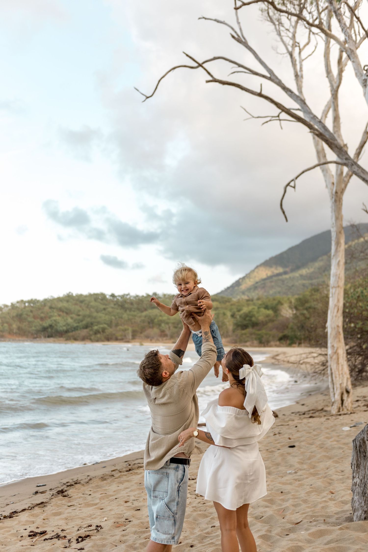 Father lifting smiling toddler high on beach while mother stands beside bare coastal trees.