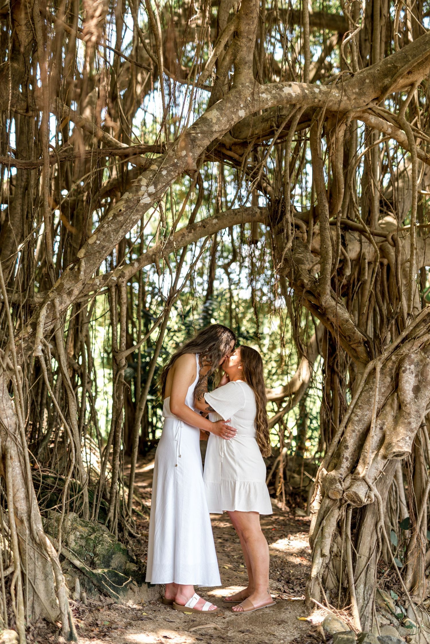 Two women in white dresses share a romantic kiss beneath a natural archway of banyan tree roots in a lush forest.