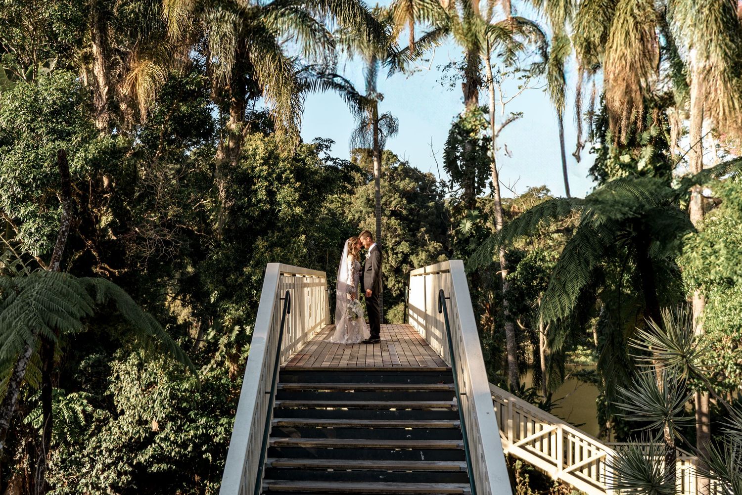Bride and groom share a romantic moment on a wooden bridge surrounded by lush tropical palm trees.