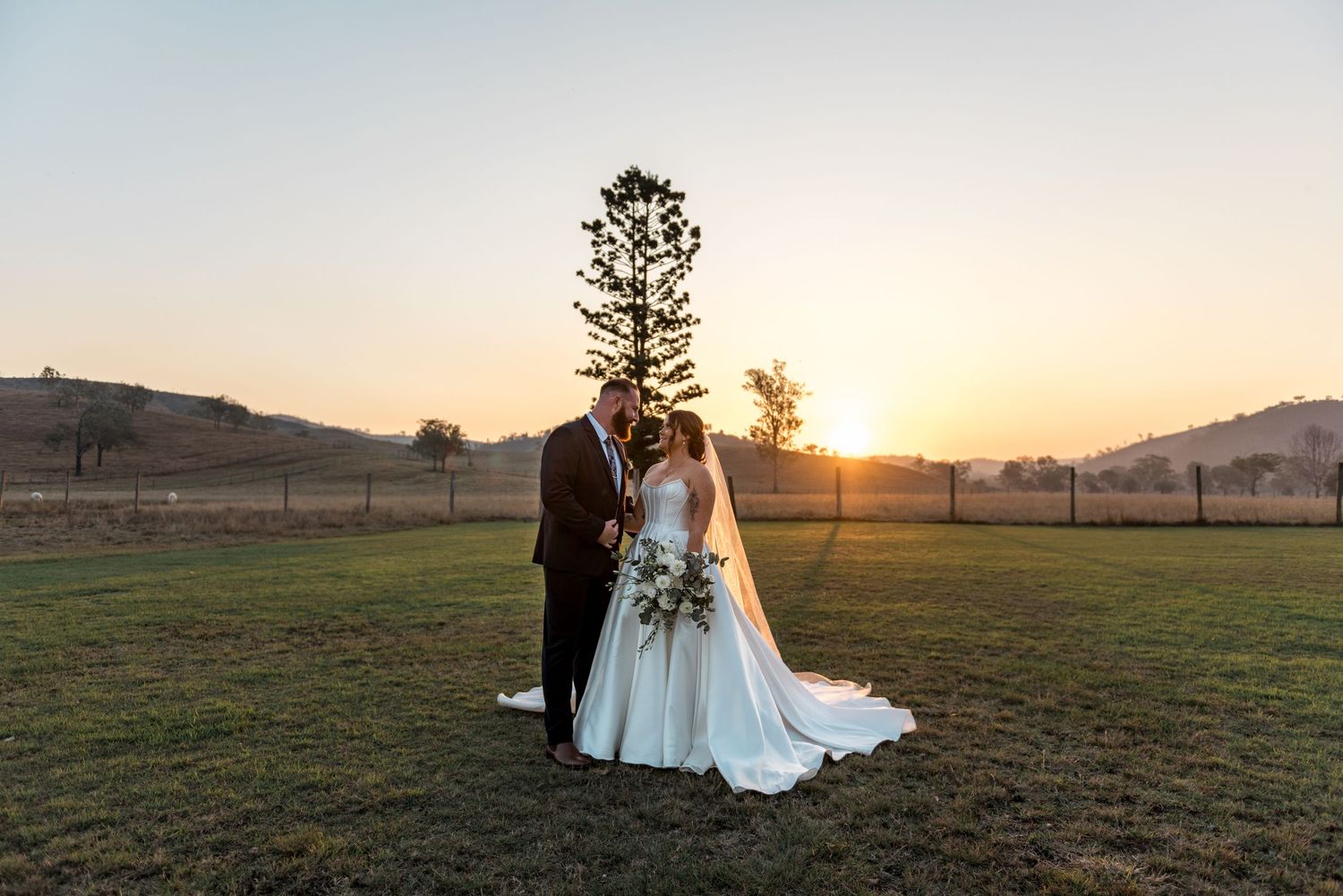 Bride and groom standing close together at golden sunset with pine tree and rolling hills behind.