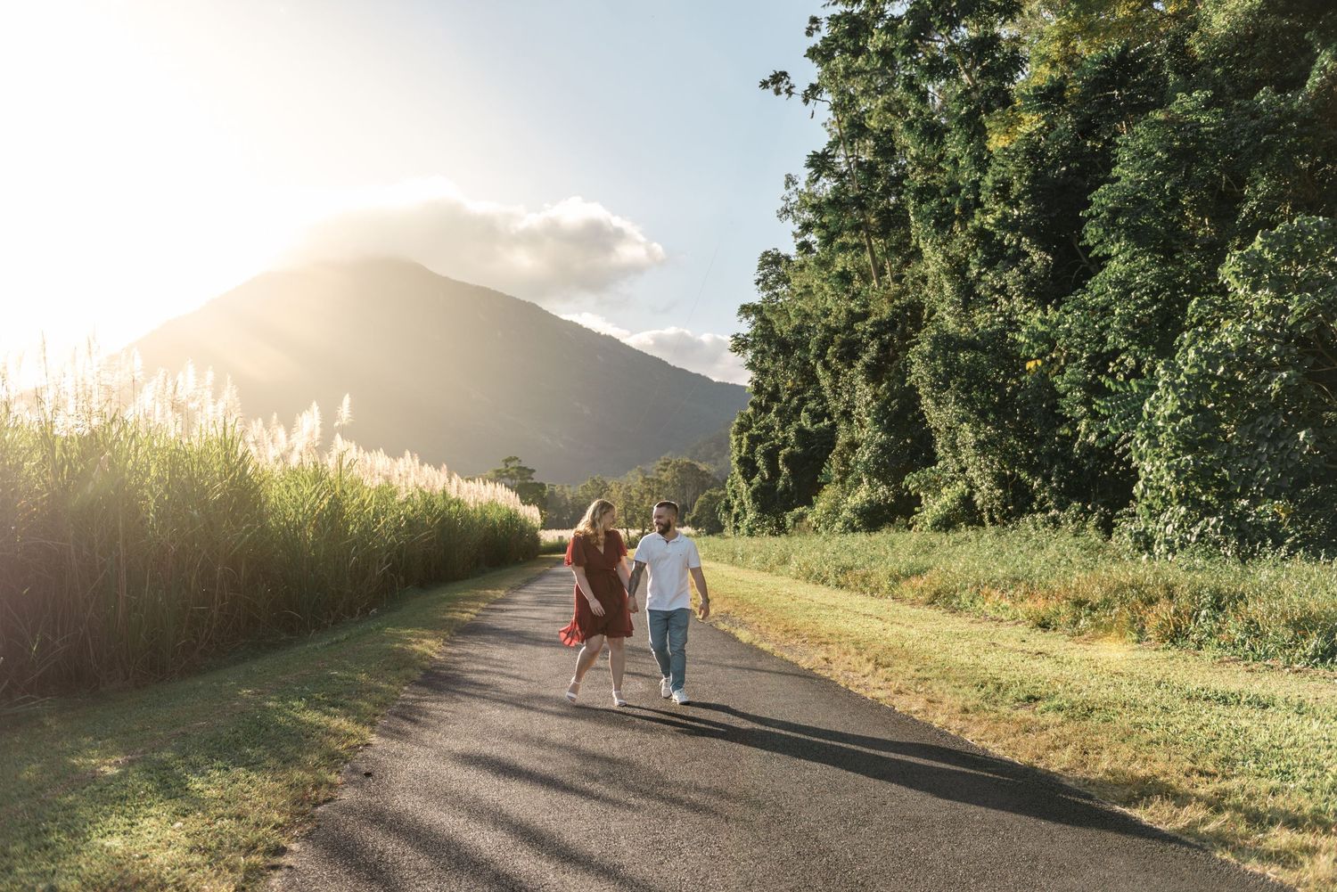 Couple holding hands walking away on a country road surrounded by tall grass and mountains at golden hour.