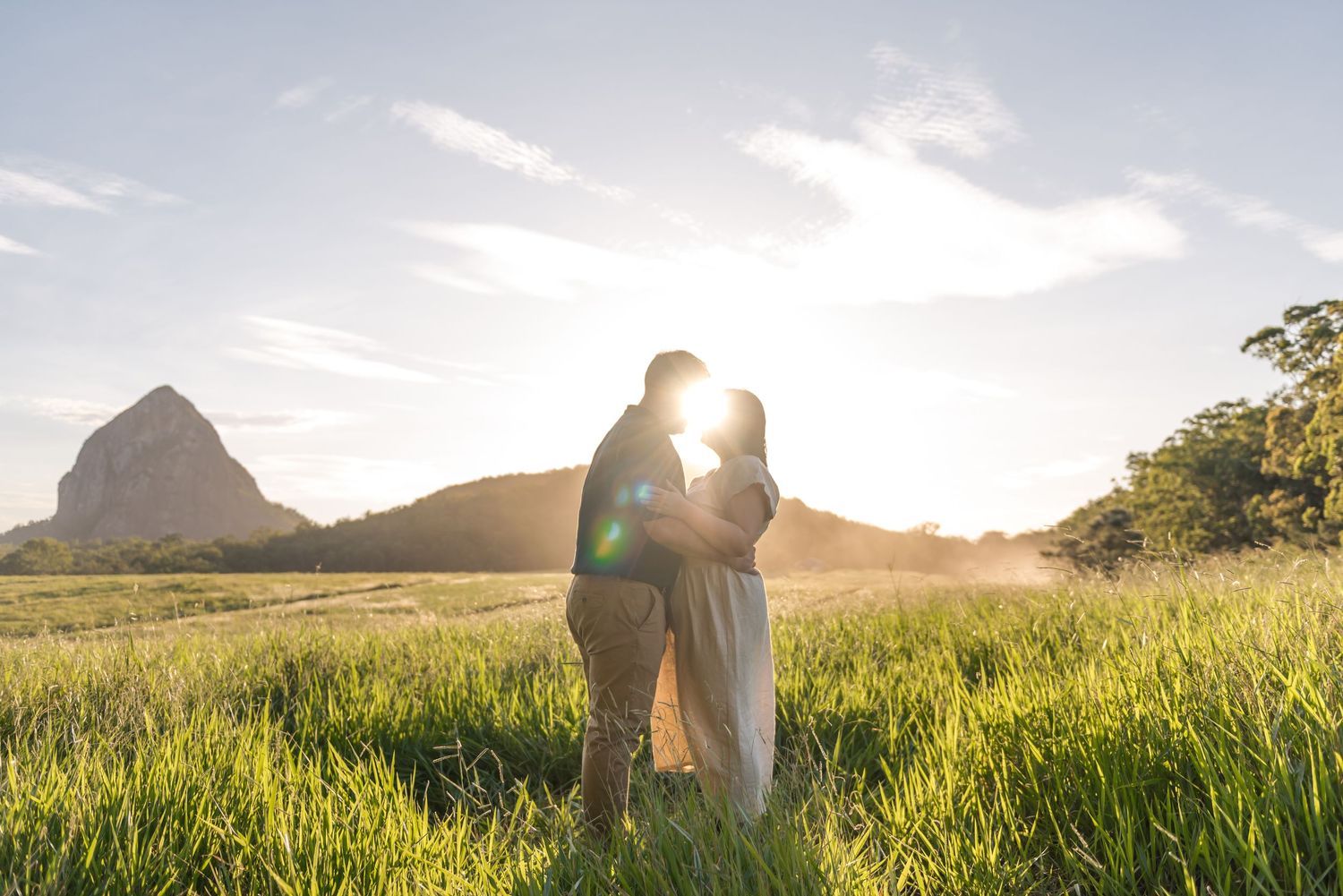 Couple sharing romantic kiss silhouetted against glowing sunset with mountain landscape behind.
